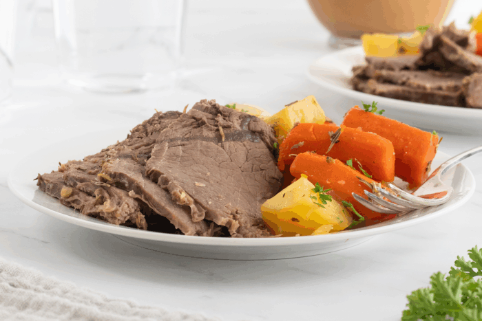 A plate of sliced pot roast served with chunks of carrots and potatoes, garnished with fresh herbs, on a white table with a fork beside the food.