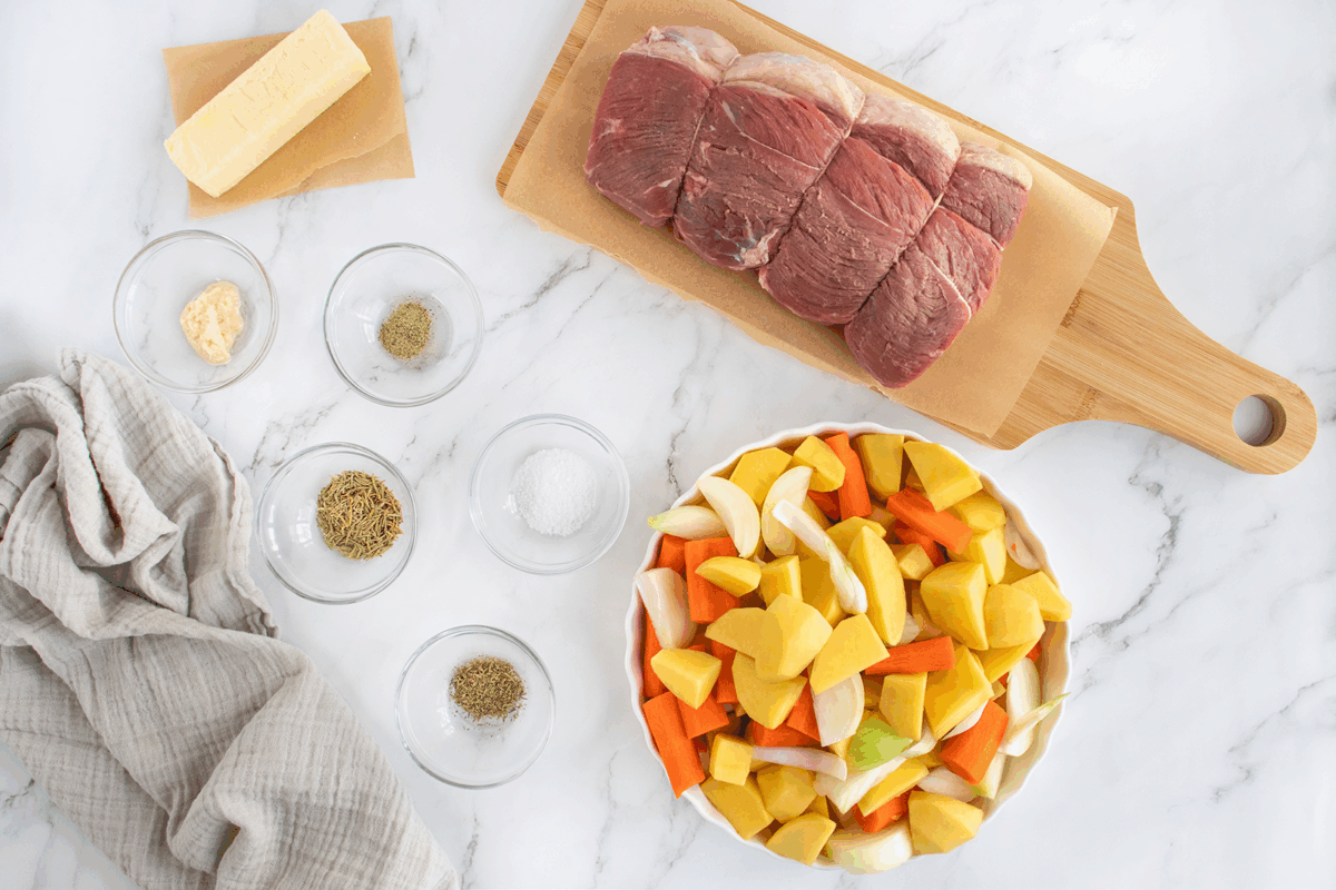Raw beef roast on a wooden board, a bowl of chopped potatoes, carrots, and onions, butter, garlic, spices, and a beige cloth on a white marble surface.