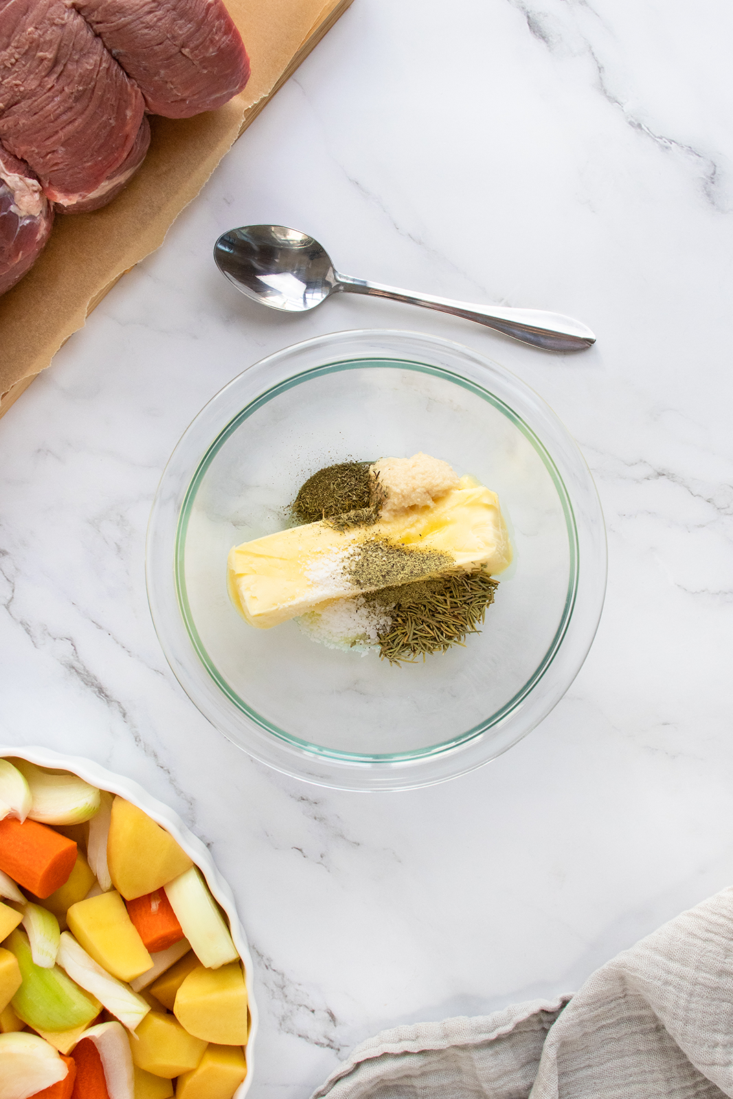 A glass bowl with a stick of butter and various herbs and seasonings on a marble countertop. Nearby are a metal spoon, raw beef, and a bowl of chopped vegetables.