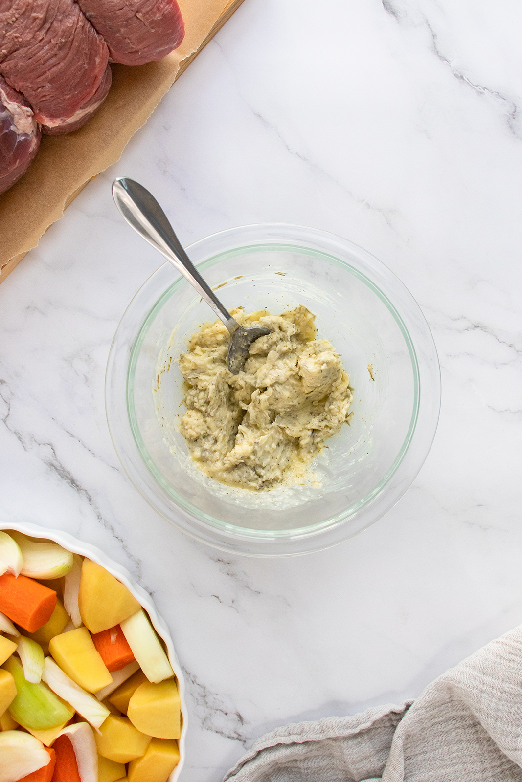 A glass bowl with a mashed mixture and a fork sits on a marble counter next to a tray of raw meat and a dish of chopped root vegetables. A striped cloth is partially visible in the corner.
