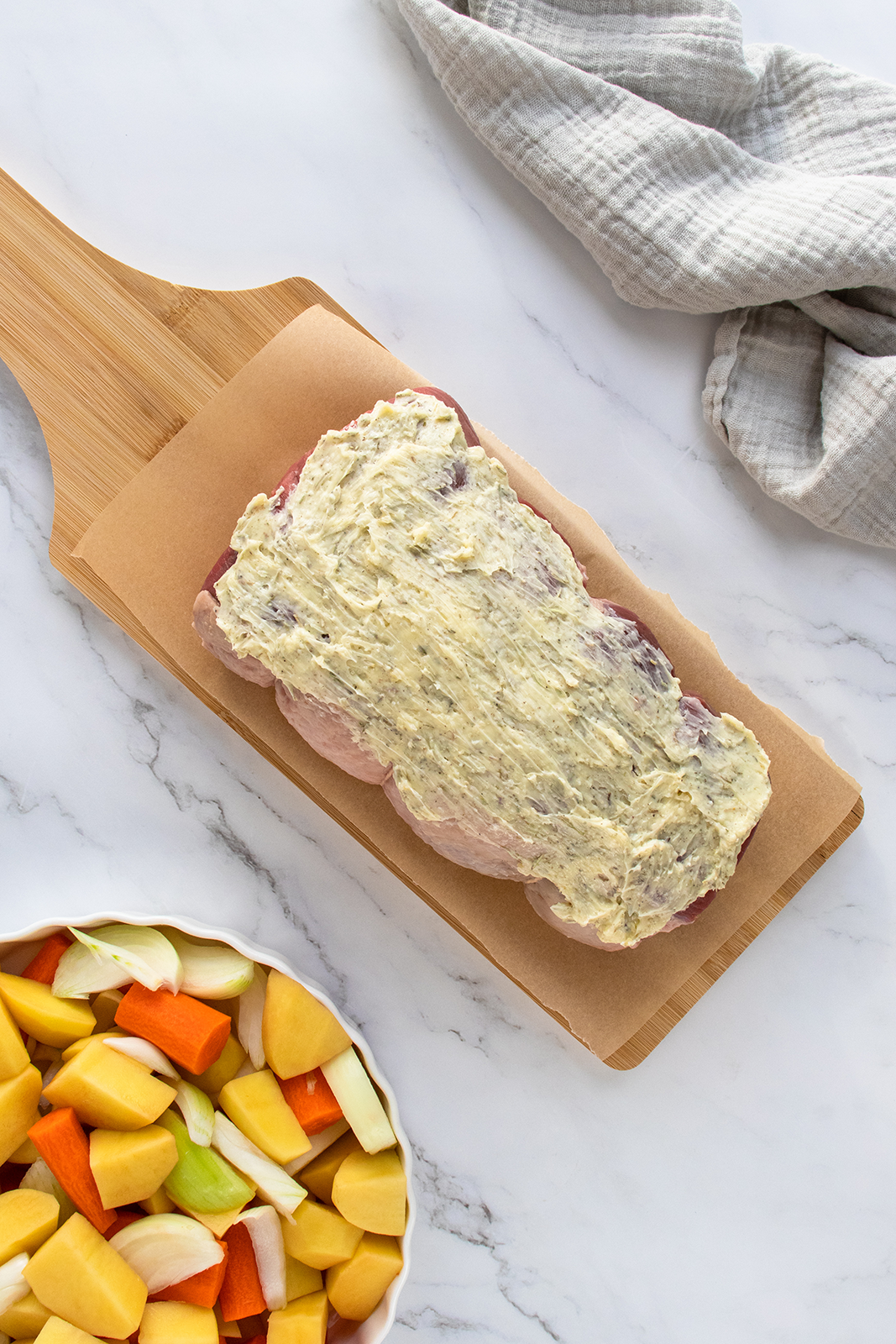 A raw pork loin coated with an herb butter mixture sits on a wooden cutting board lined with parchment paper. Nearby is a bowl of chopped vegetables and a folded gray kitchen towel on a marble countertop.