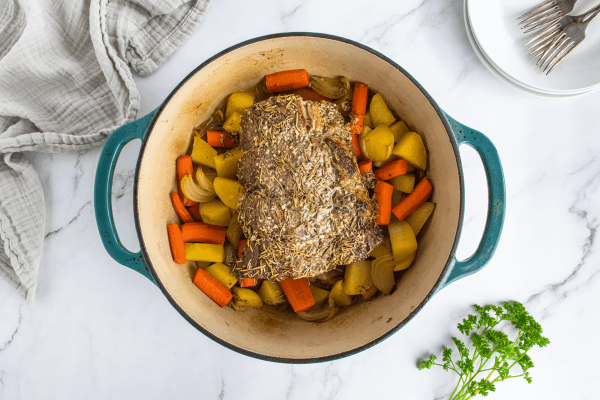 A cooked pot roast with carrots, potatoes, and onions sits in a teal Dutch oven on a marble countertop, with a beige cloth napkin, parsley, plates, and forks nearby.
