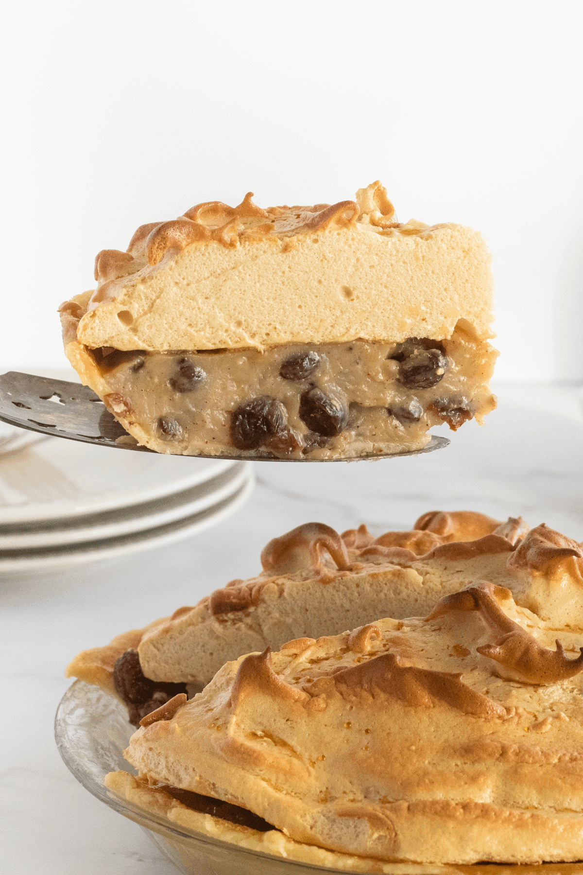 A slice of pie with a golden-brown meringue topping and a creamy filling with visible raisins is being lifted from a whole pie on a glass dish. Plates are stacked in the background.