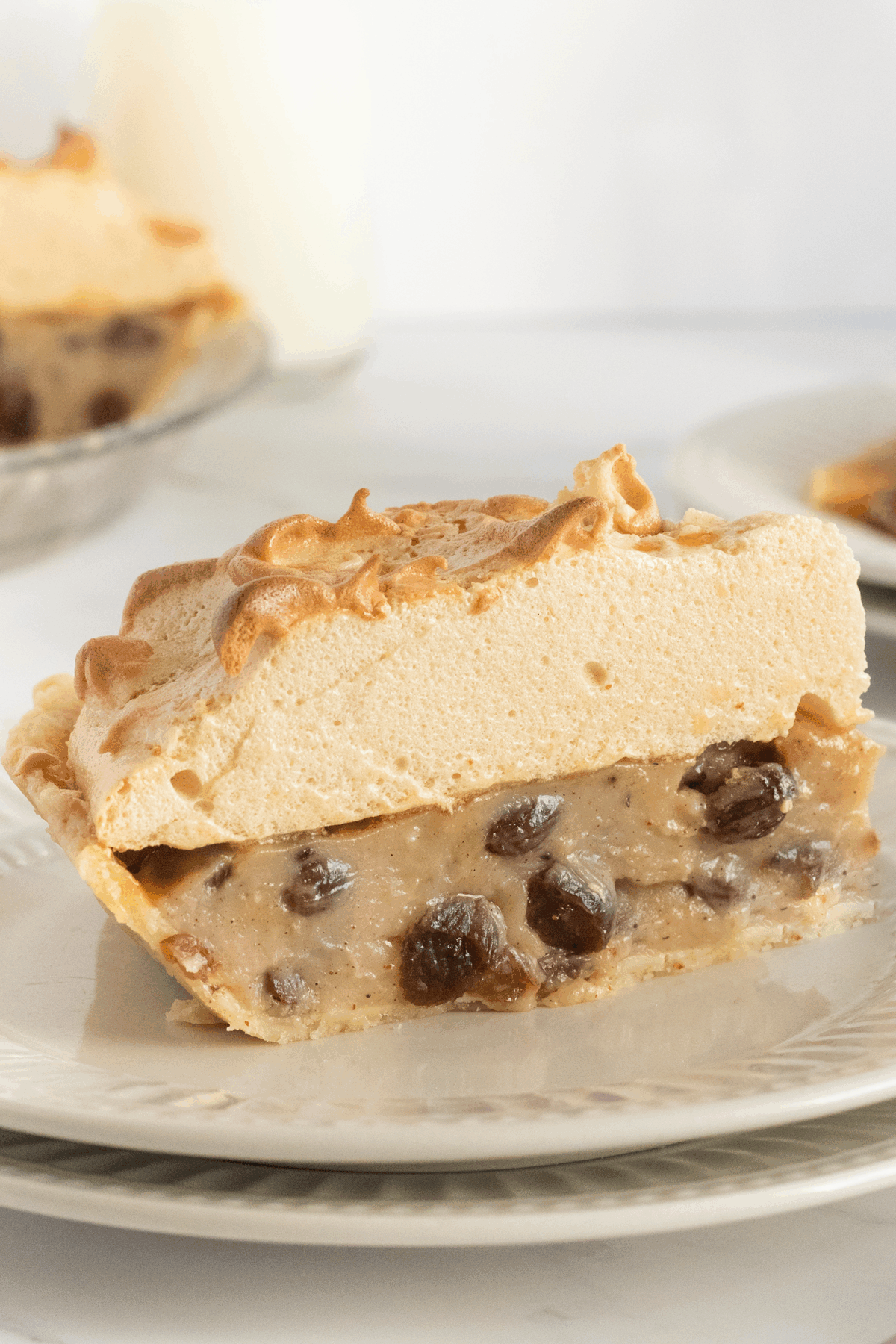 A slice of pie with a thick, golden meringue topping, creamy filling with visible raisins, served on a white plate. Another slice and a glass of milk are blurred in the background.