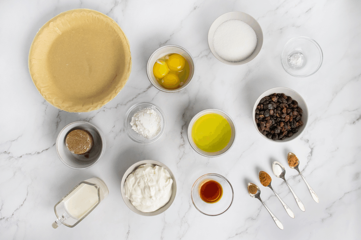 Top view of baking ingredients on a marble surface, including a pie crust, eggs, sugar, raisins, sour cream, spices on spoons, butter, vanilla, brown sugar, milk, and cornstarch, all arranged in separate bowls.