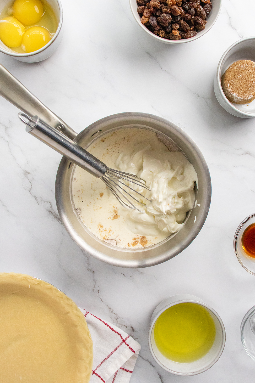 A top-down view of baking ingredients on a marble surface, including a pie crust, egg yolks, whipped cream in a saucepan with a whisk, melted butter, vanilla extract, raisins, and a small bowl of brown sugar.