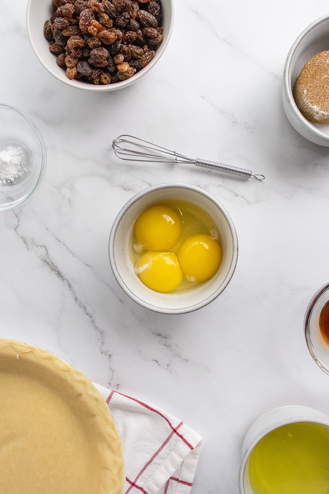 A marble countertop with baking ingredients: a pie crust, a bowl with two cracked eggs, a bowl of raisins, sugar, a small whisk, a bowl of oil, a bowl with liquid, and a bowl with baking powder.