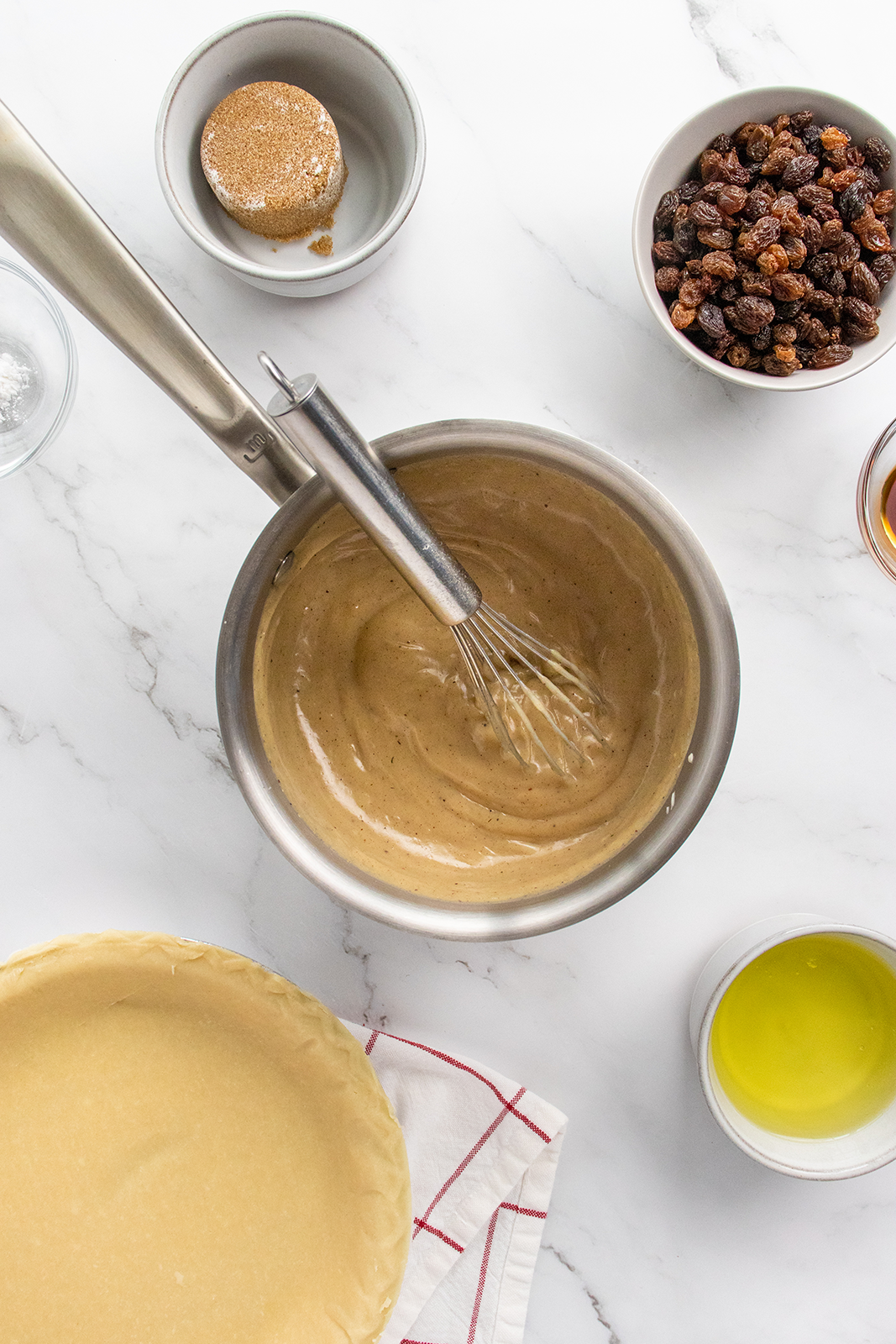 A metal bowl with a whisk mixing a tan batter sits on a marble counter, surrounded by a pie crust, bowl of raisins, brown sugar, melted butter, and a red-striped towel.