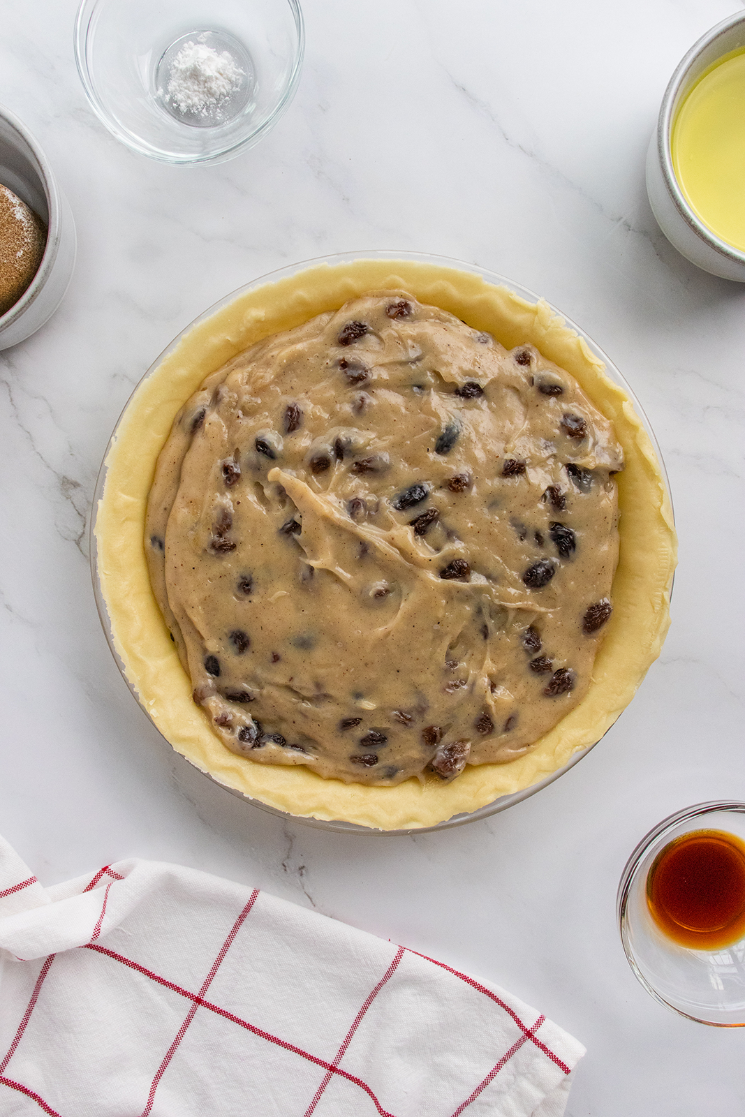 A pie crust filled with a creamy mixture containing raisins sits on a marble countertop, surrounded by baking ingredients and a white towel with red stripes.