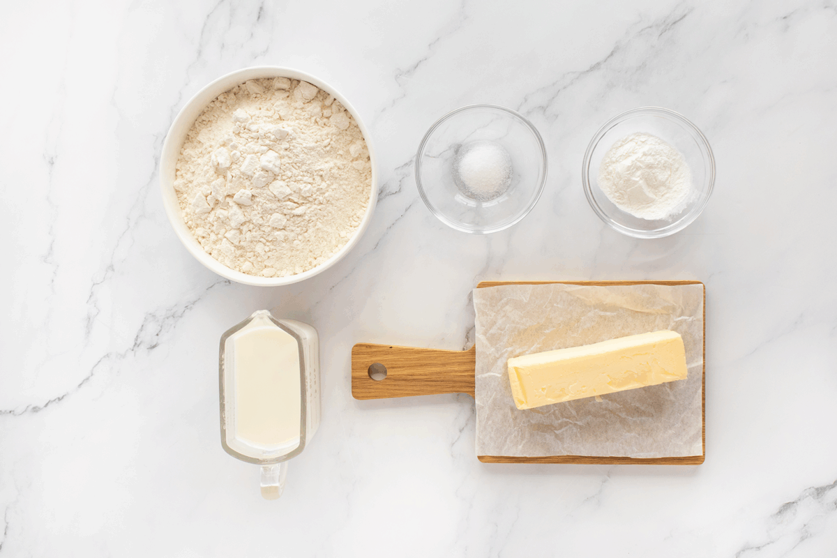 A marble surface with bowls of flour, salt, and baking powder, a glass measuring cup of milk, and a stick of butter on parchment paper atop a wooden cutting board.