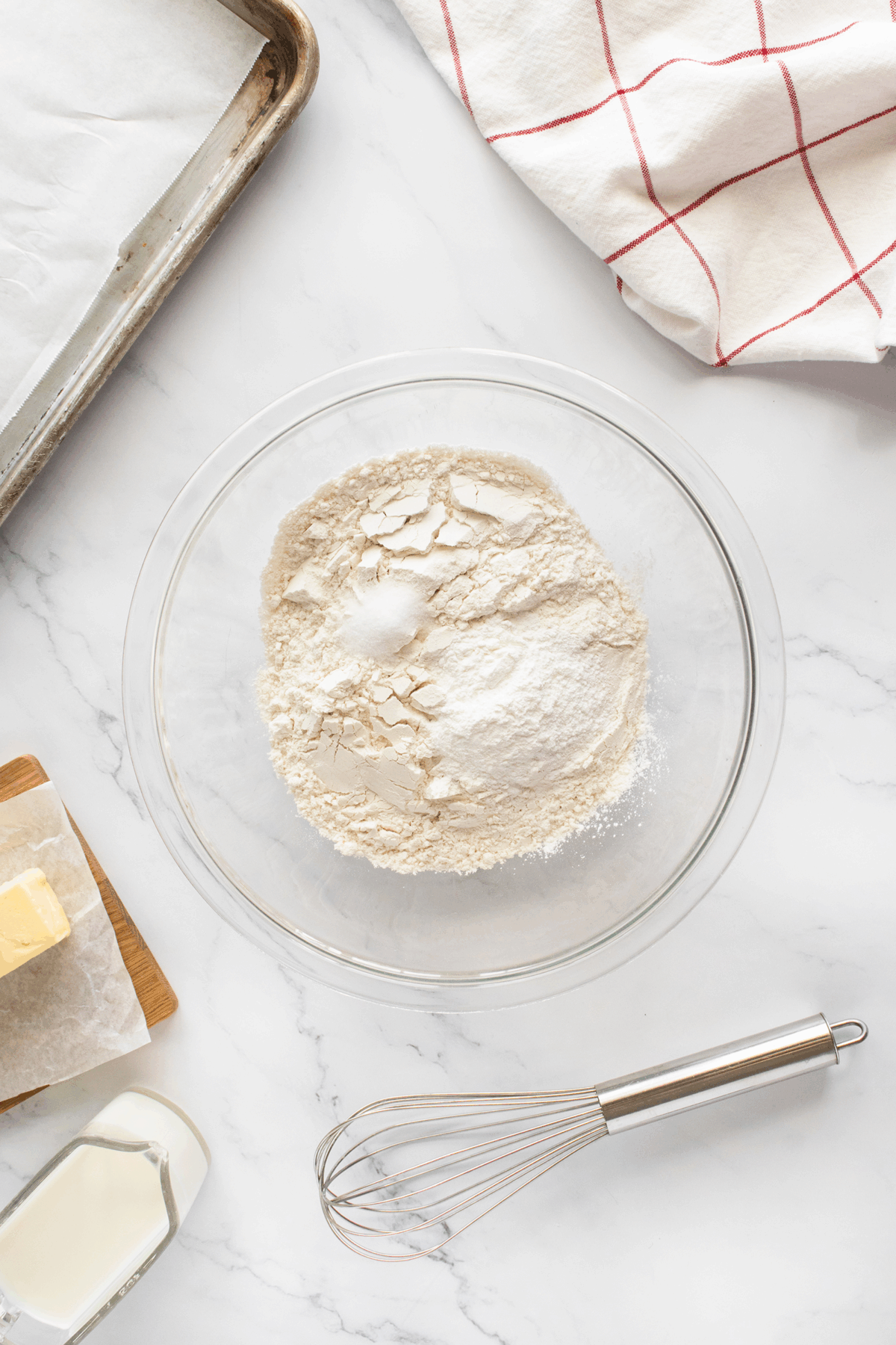 A glass bowl with flour, sugar, and baking powder sits on a white countertop near a metal whisk, butter, milk, a baking tray with parchment, and a red-and-white kitchen towel.