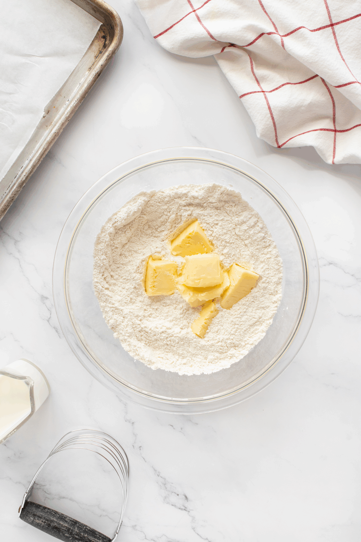 A glass bowl with flour and chunks of butter sits on a marble countertop, surrounded by a baking sheet, a kitchen towel, a metal pastry cutter, and a bottle of milk.
