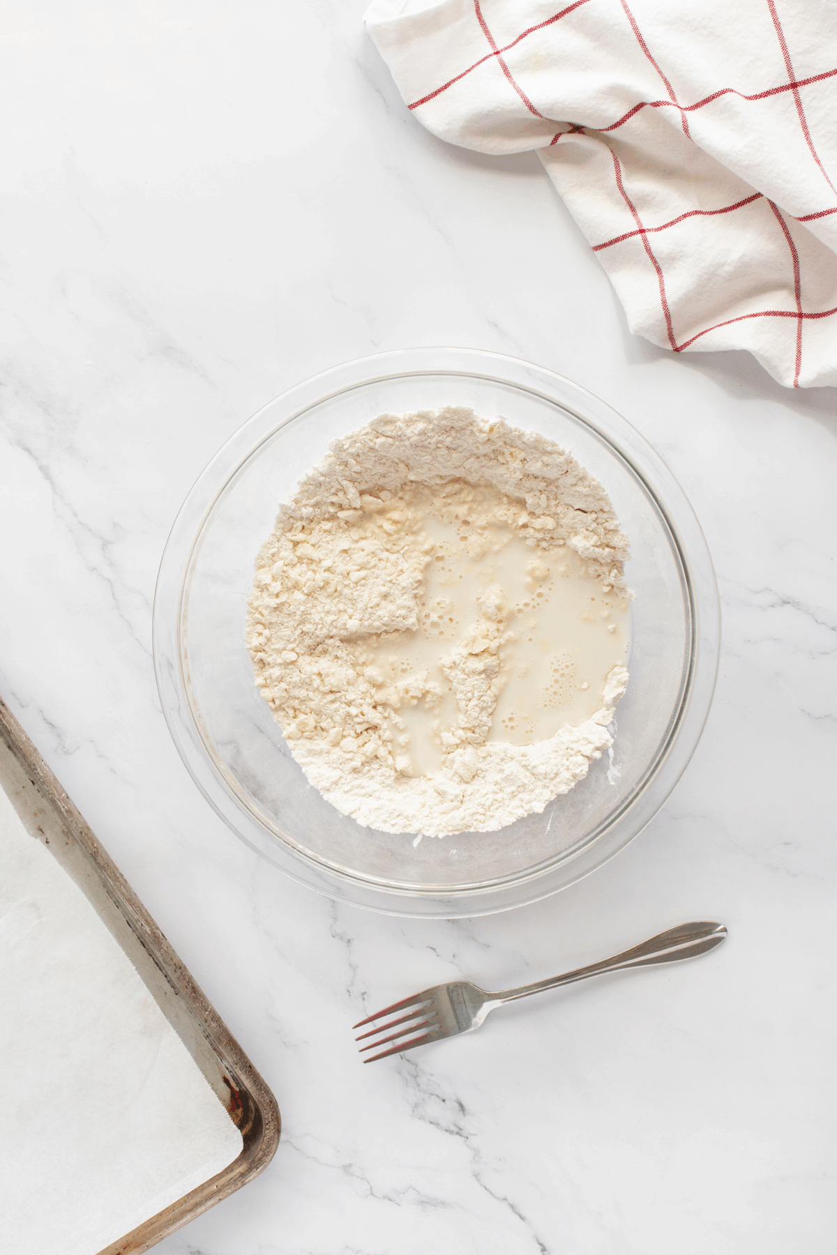 A glass bowl with flour and milk sits on a marble surface next to a fork, a parchment-lined baking sheet, and a white towel with red stripes.