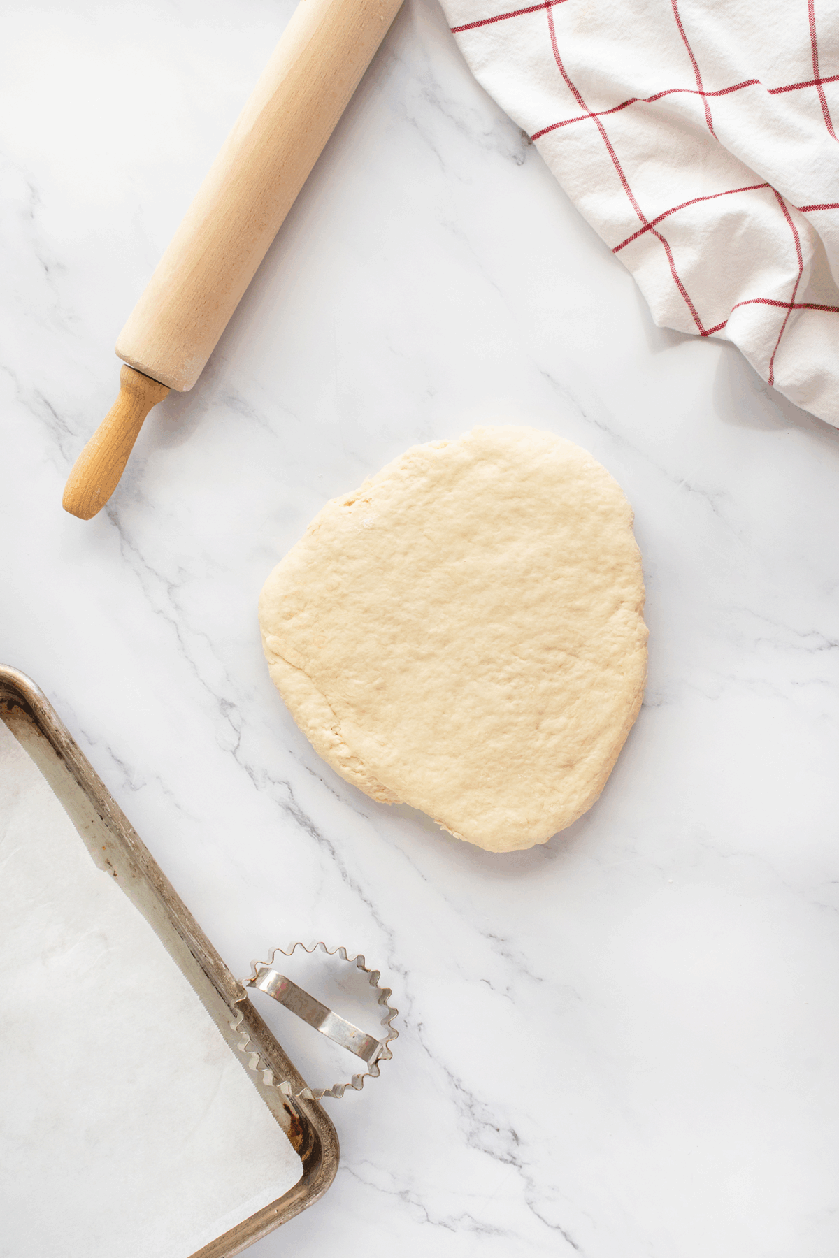 A ball of dough sits on a marble surface beside a rolling pin, a baking sheet lined with parchment paper, a metal biscuit cutter, and a white towel with red stripes.