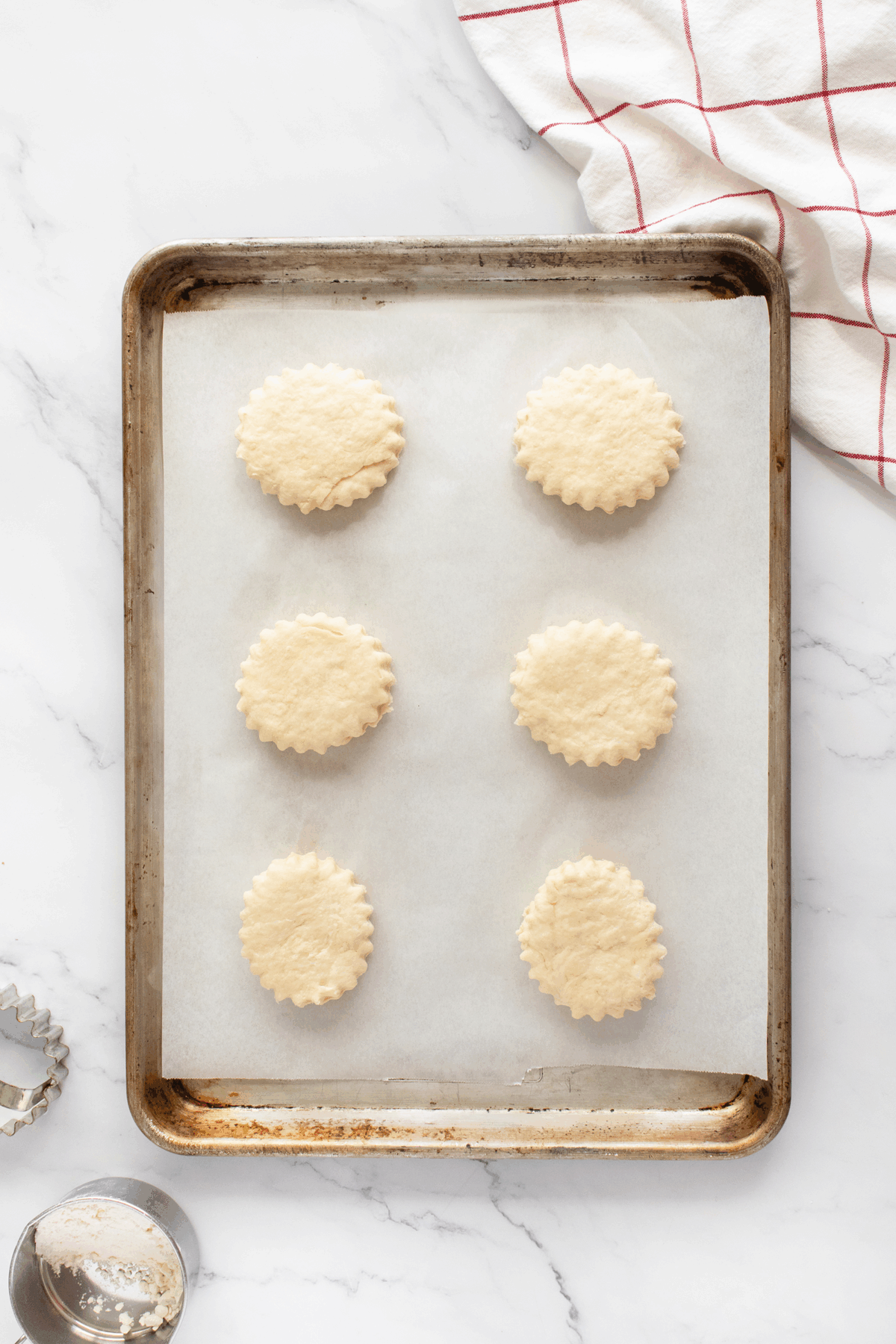 A baking sheet lined with parchment paper holds six unbaked, round, scalloped-edge cookies. A cookie cutter and a dusting of flour are nearby on a white marble surface with a red and white kitchen towel.