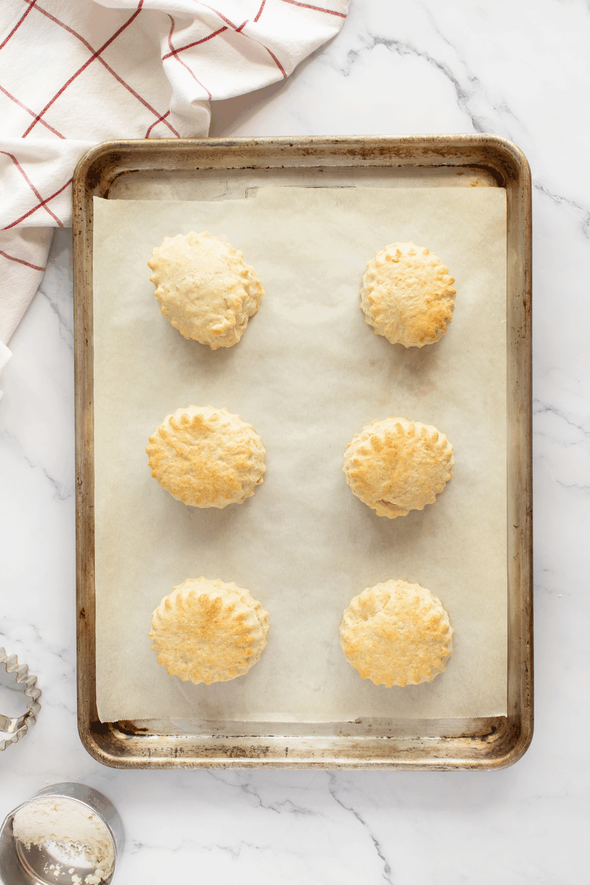 A baking sheet lined with parchment paper holds six golden brown biscuits. A red and white checkered towel is in the corner, next to a biscuit cutter and a bowl with flour on a marble surface.