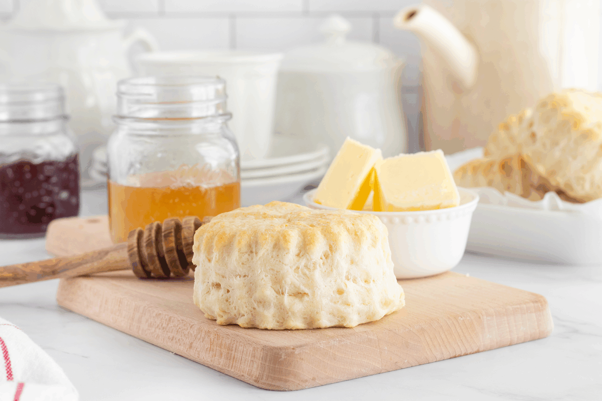 A freshly baked biscuit sits on a wooden board next to a honey dipper, a jar of honey, a small bowl of butter, and stacked biscuits in the background on a white kitchen counter.