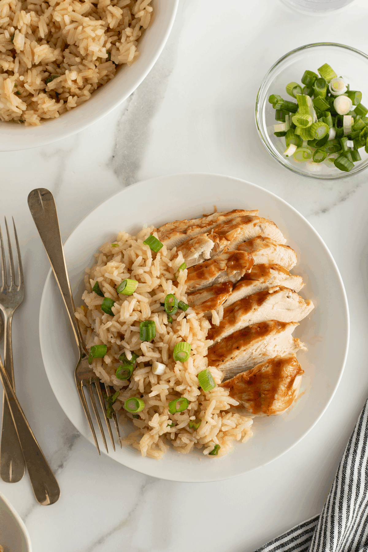 A white plate with sliced grilled chicken breast and rice topped with chopped green onions; a fork and knife are on the side. A bowl of rice and a small bowl of green onions are nearby on a marble surface.