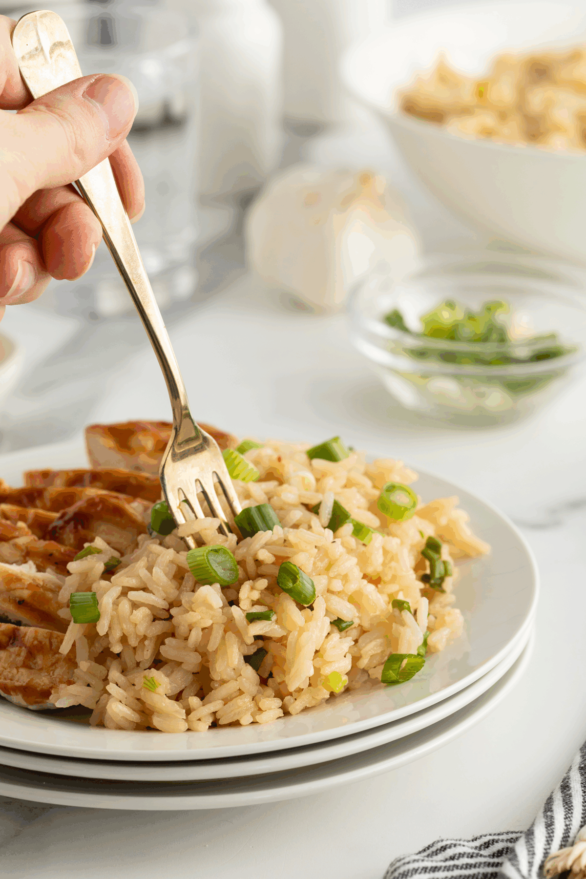 A hand holds a fork above a plate of rice with chopped green onions and grilled chicken slices. In the background are a bowl of more rice, a glass of water, and a bowl of chopped green onions.