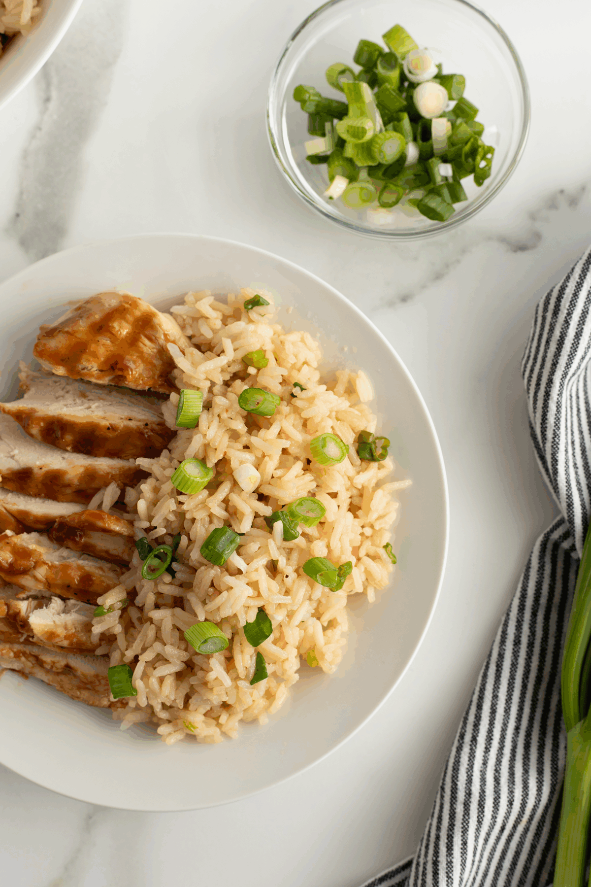 A plate with sliced grilled chicken and rice topped with chopped green onions. Nearby, a bowl of sliced green onions and a striped cloth napkin rest on a white marble surface.