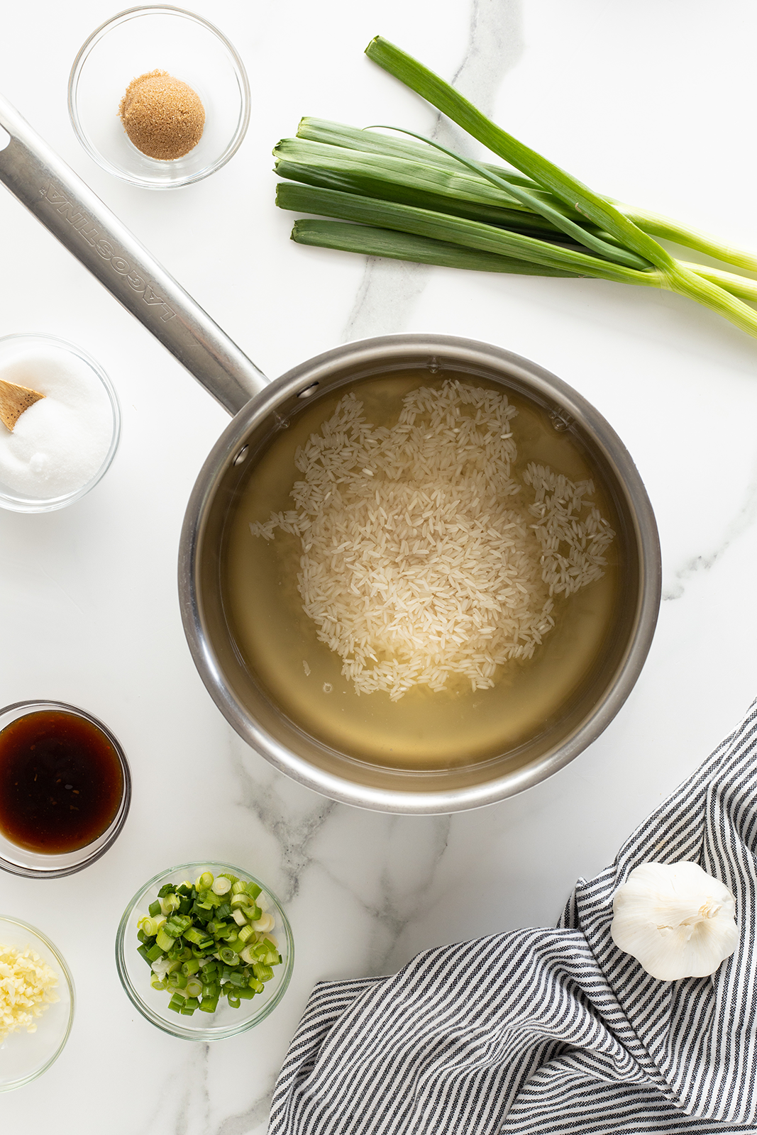 A saucepan with rice and water sits on a white surface surrounded by bowls of brown sugar, white sugar, soy sauce, chopped green onions, minced garlic, and whole green onions. A striped cloth and a garlic bulb are nearby.