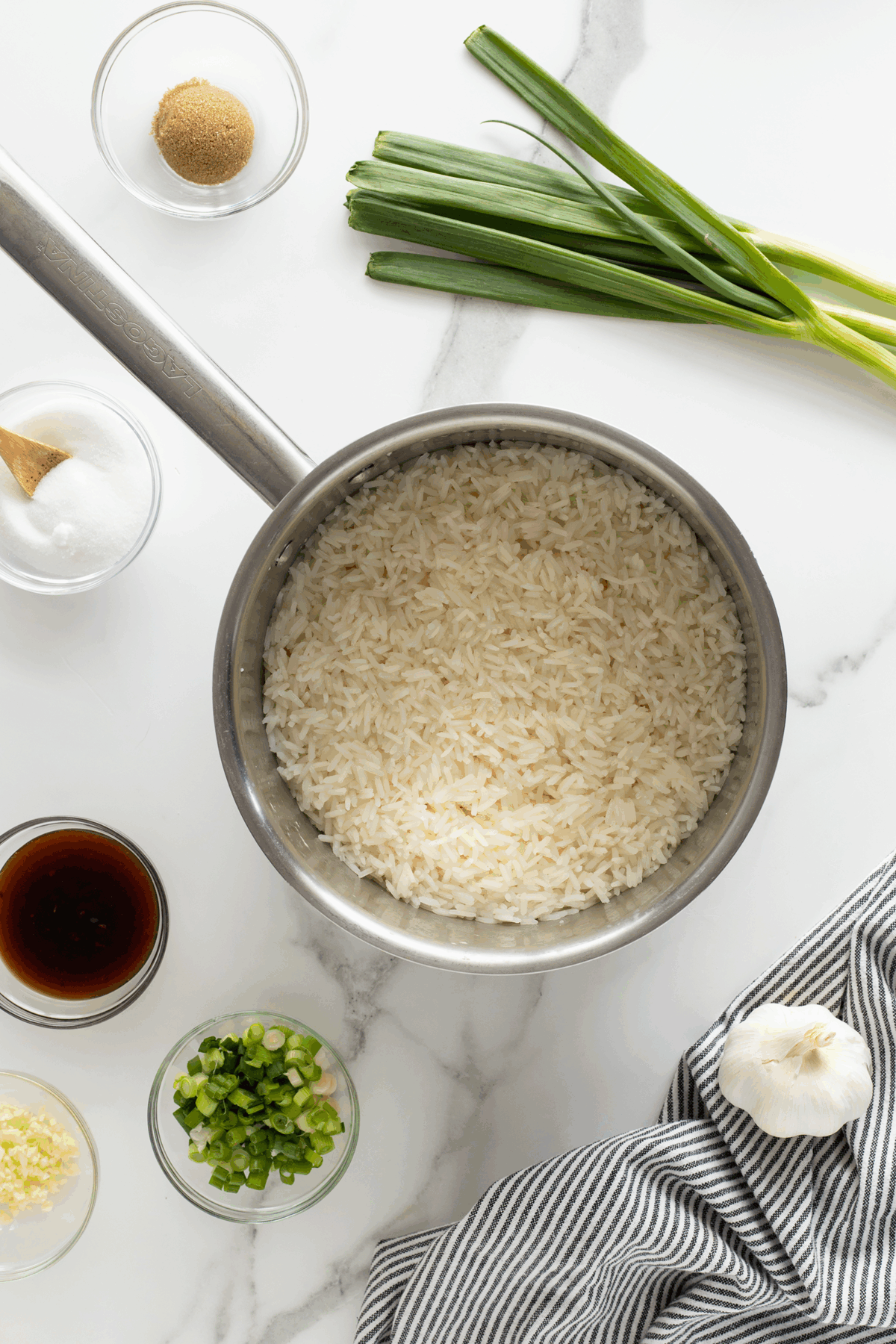 A saucepan filled with cooked white rice sits on a marble counter, surrounded by green onions, garlic, chopped scallions, ginger, sugar, brown sugar, and soy sauce in small bowls. A striped towel is partially visible.