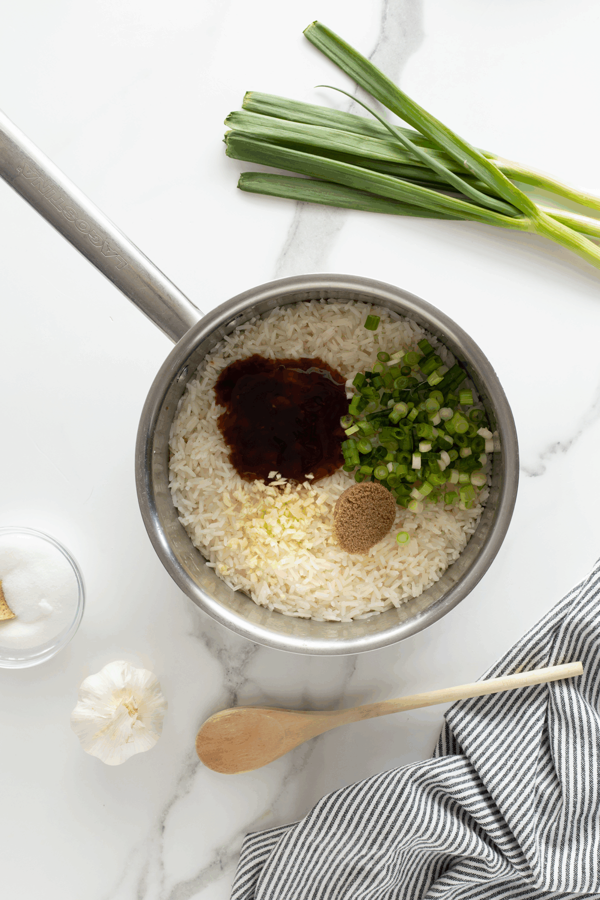 A saucepan on a marble surface contains uncooked rice, chopped green onions, minced garlic, a dark sauce, and a brown spice. Nearby are fresh green onions, a garlic bulb, a bowl of sugar, a wooden spoon, and a striped cloth.