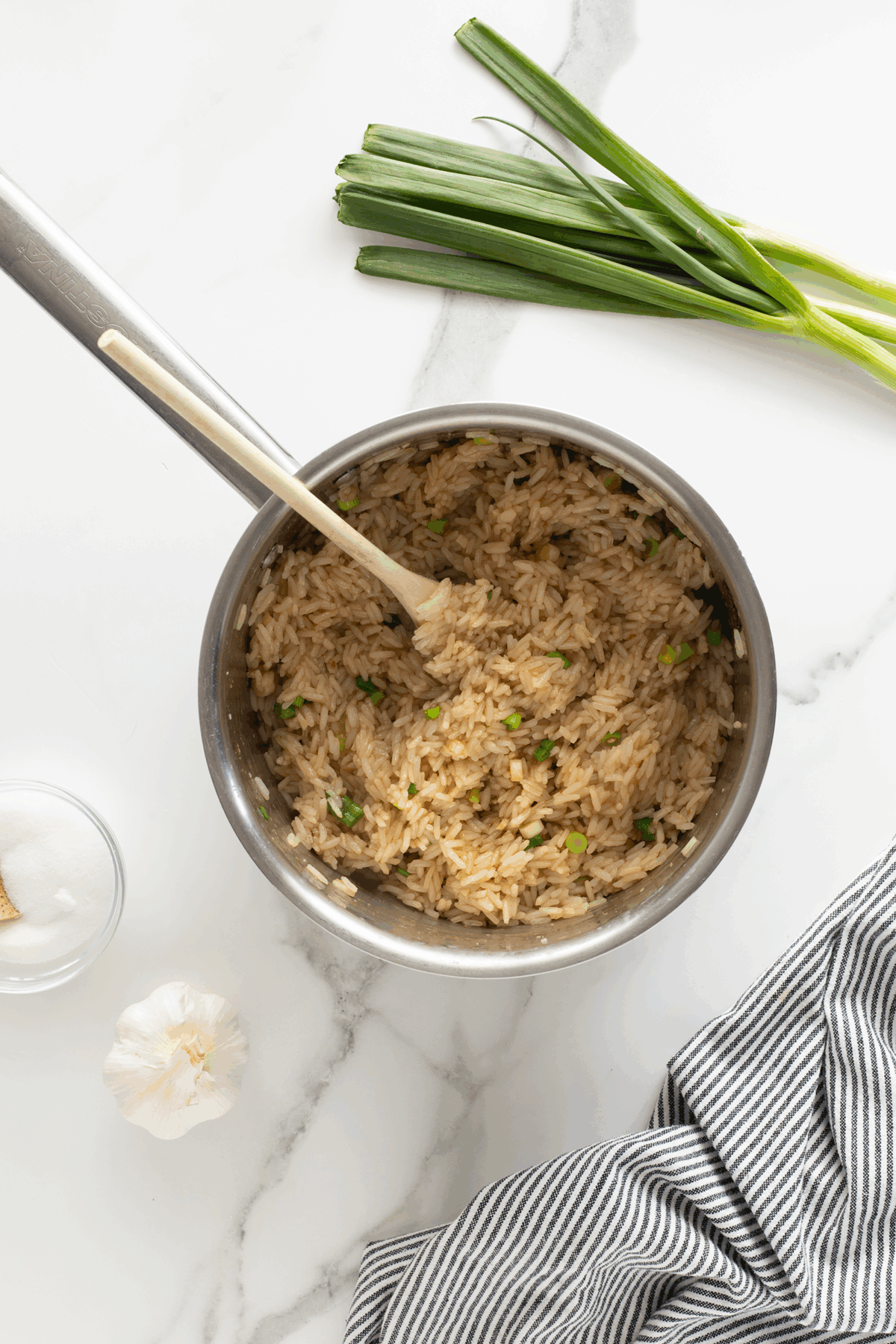 A pot of cooked rice mixed with green onions and a wooden spoon, placed on a white marble countertop. Nearby are fresh green onions, a garlic bulb, a small bowl, and a striped cloth napkin.
