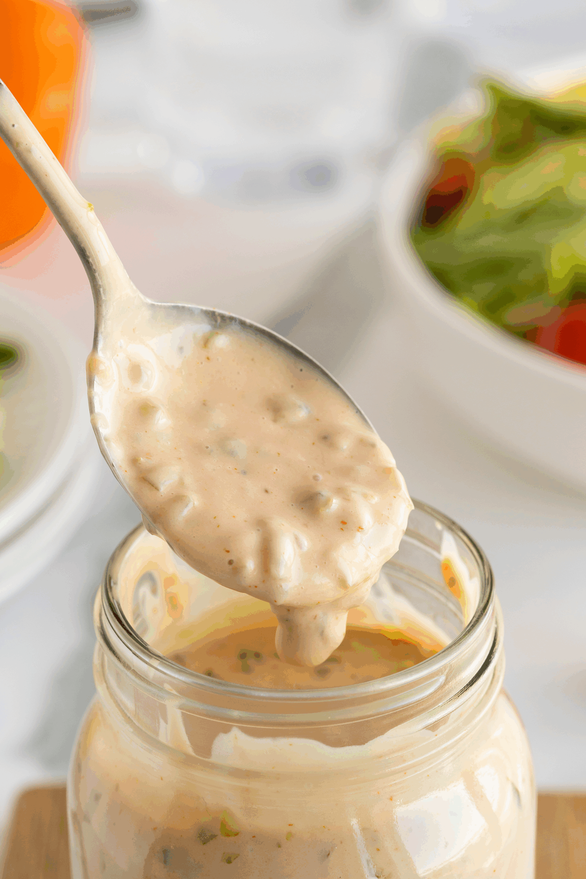 A spoonful of creamy, chunky salad dressing is held above an open glass jar, with a blurred bowl of salad in the background.