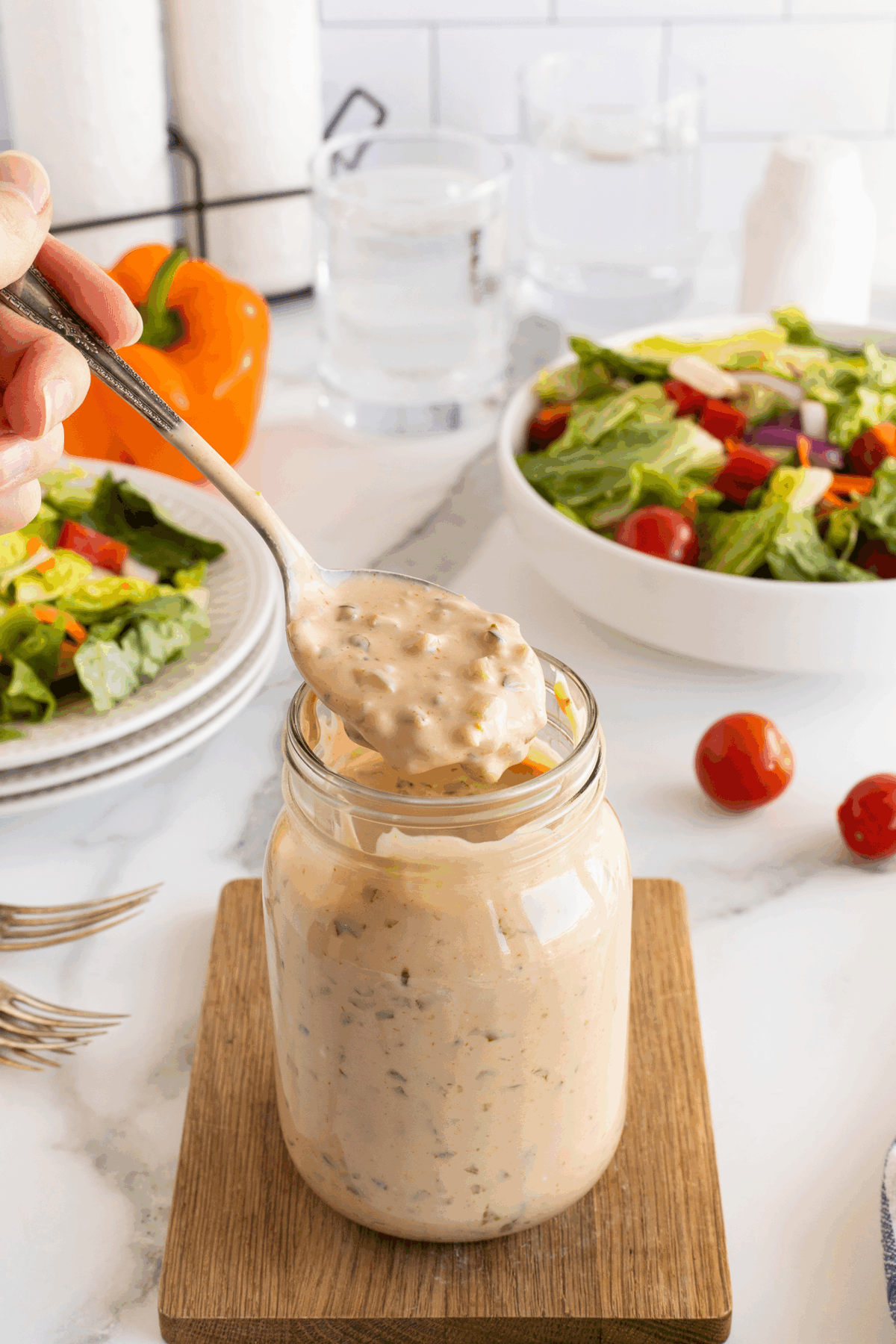 A spoonful of creamy dressing is held above a glass jar, with a fresh salad, tomatoes, and a bell pepper in the background on a kitchen counter.
