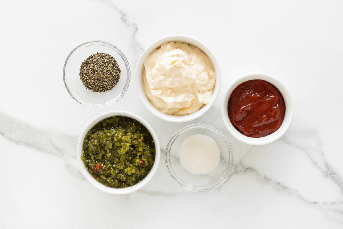 Five small bowls on a marble surface containing black pepper, mayonnaise, ketchup, chopped pickles or relish, and granulated sugar, arranged in a neat cluster.