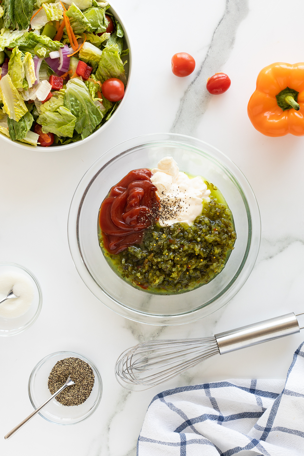 A glass bowl with mayonnaise, ketchup, relish, and pepper sits beside a whisk, a bowl of salad, a small bowl of pepper, a yellow bell pepper, grape tomatoes, and a striped kitchen towel on a white countertop.