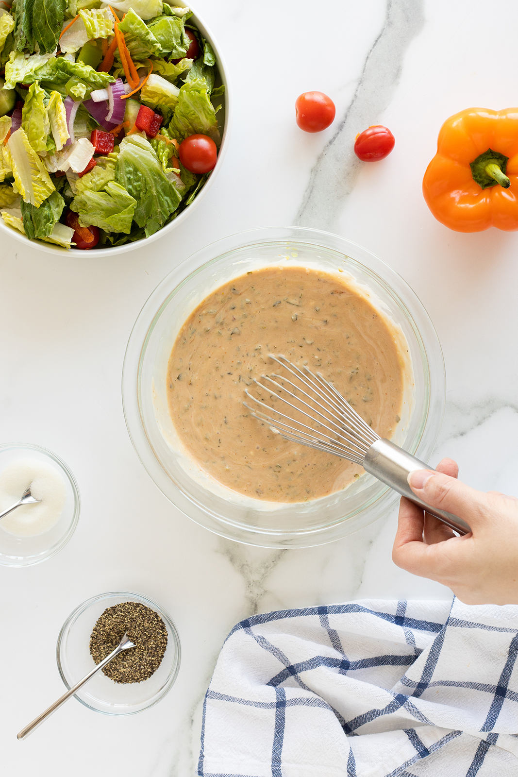 A hand whisking creamy salad dressing in a glass bowl, surrounded by a bowl of fresh salad, a small bowl of ground pepper, a small bowl of white sauce, a yellow bell pepper, grape tomatoes, and a striped kitchen towel.