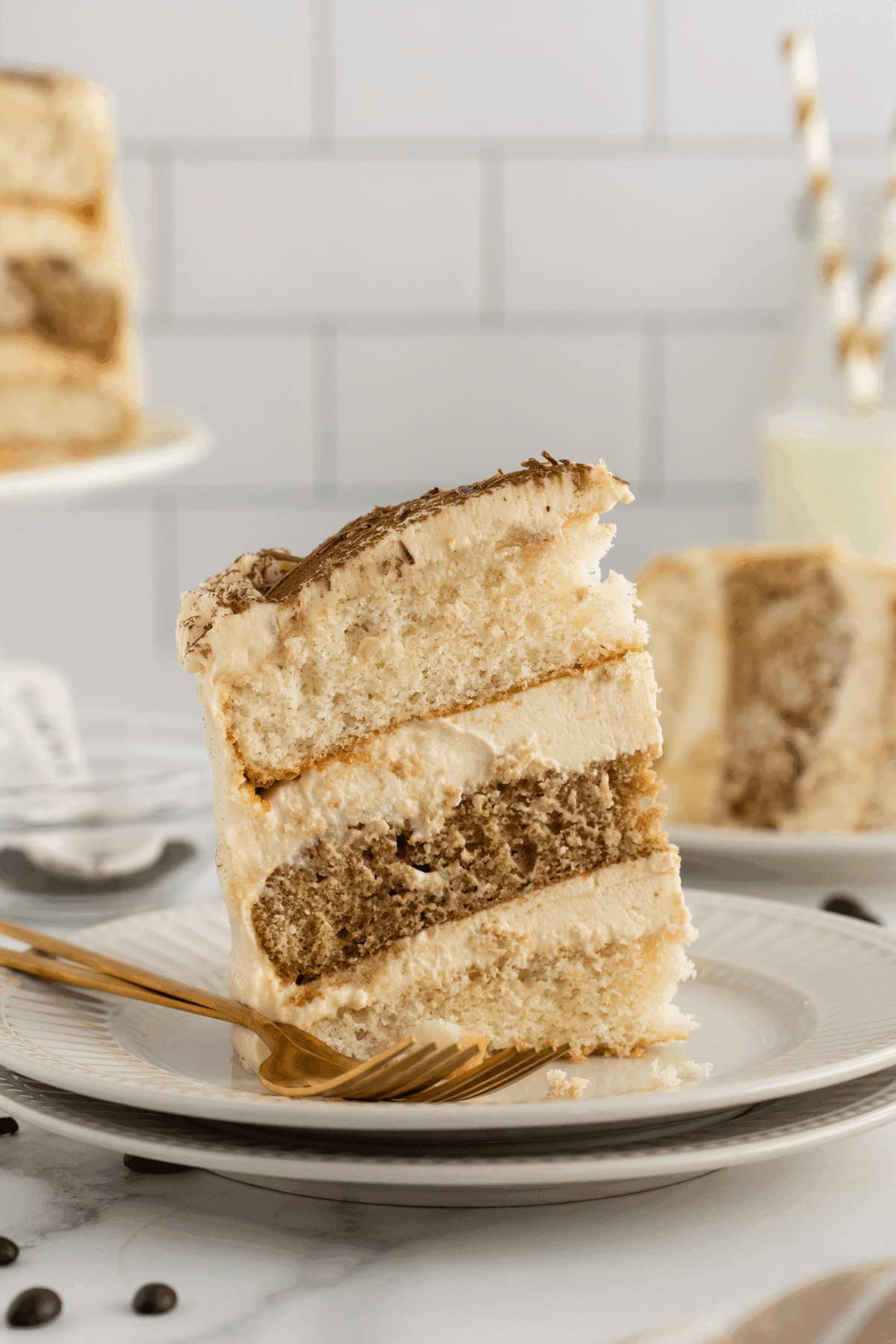 A slice of layered tiramisu cake with creamy frosting sits on a white plate with a gold fork, with a blurred background of another cake slice, a cake stand, and a bottle of milk with straws.