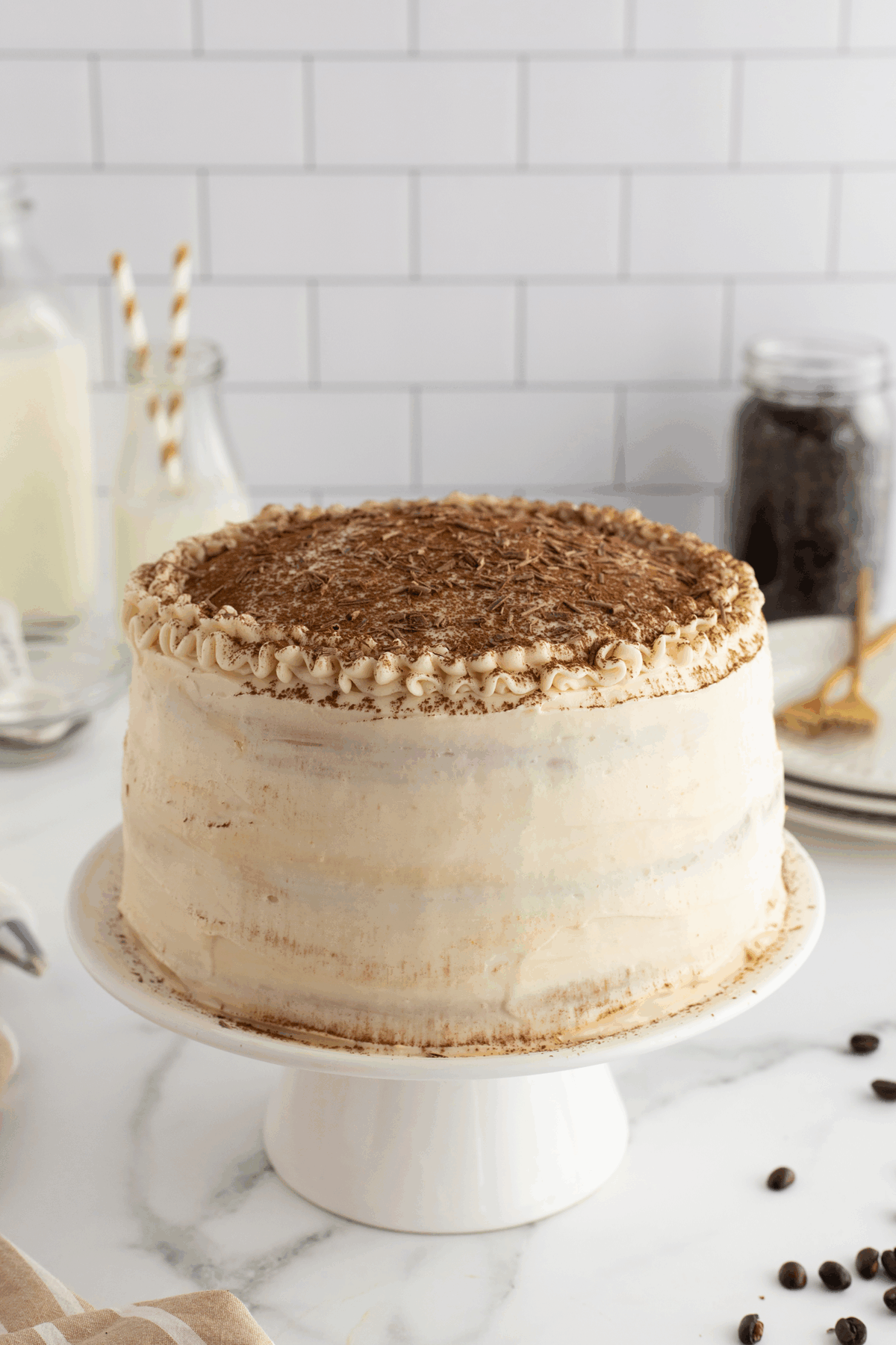 A tall layer cake with light frosting and chocolate shavings on top sits on a white cake stand. In the background are bottles of milk, a jar of coffee beans, and gold forks on a white marble counter.