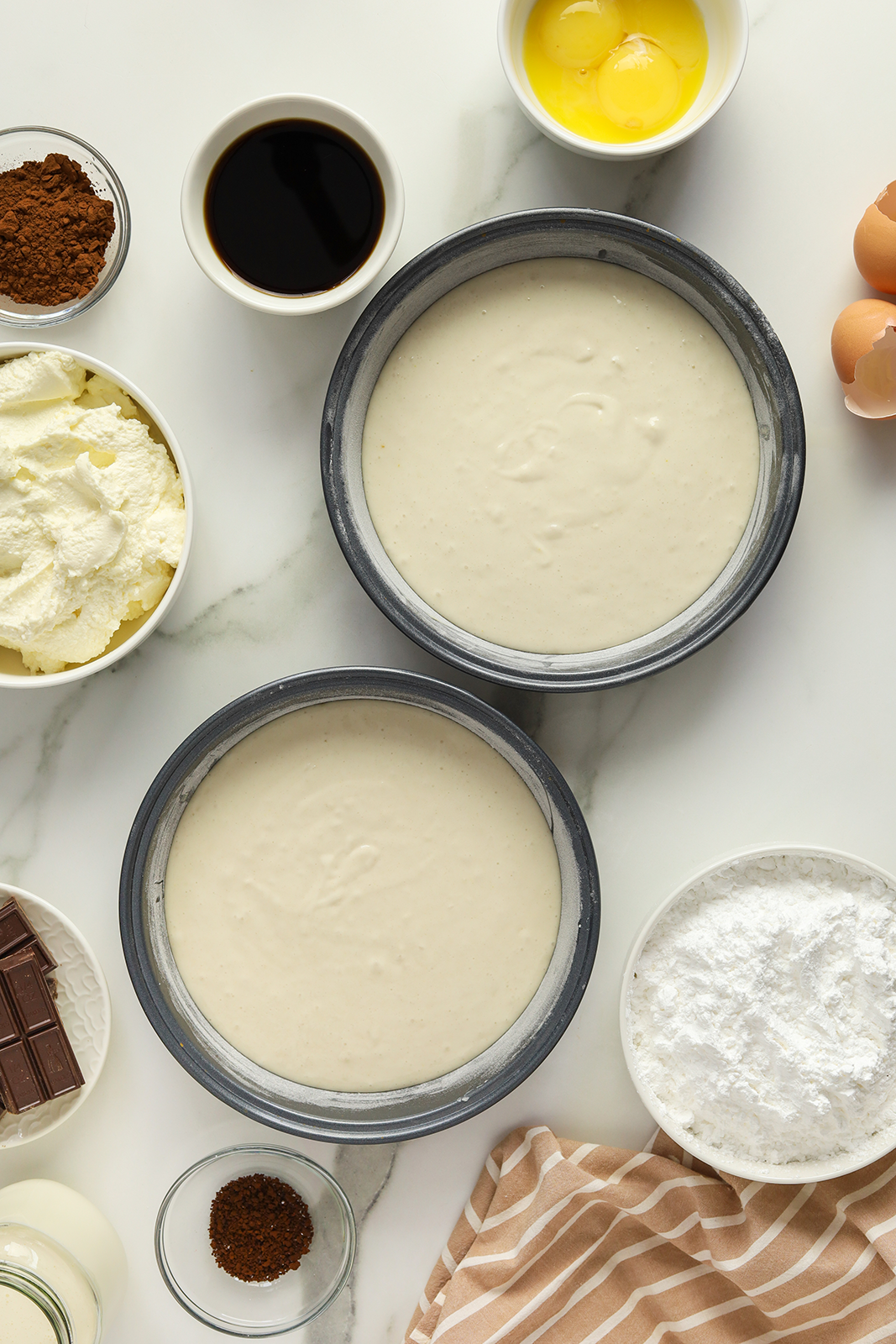Two round cake pans filled with batter are surrounded by bowls containing eggs, ricotta cheese, cocoa powder, coffee, powdered sugar, chocolate, vanilla extract, and a beige-striped kitchen towel on a white surface.