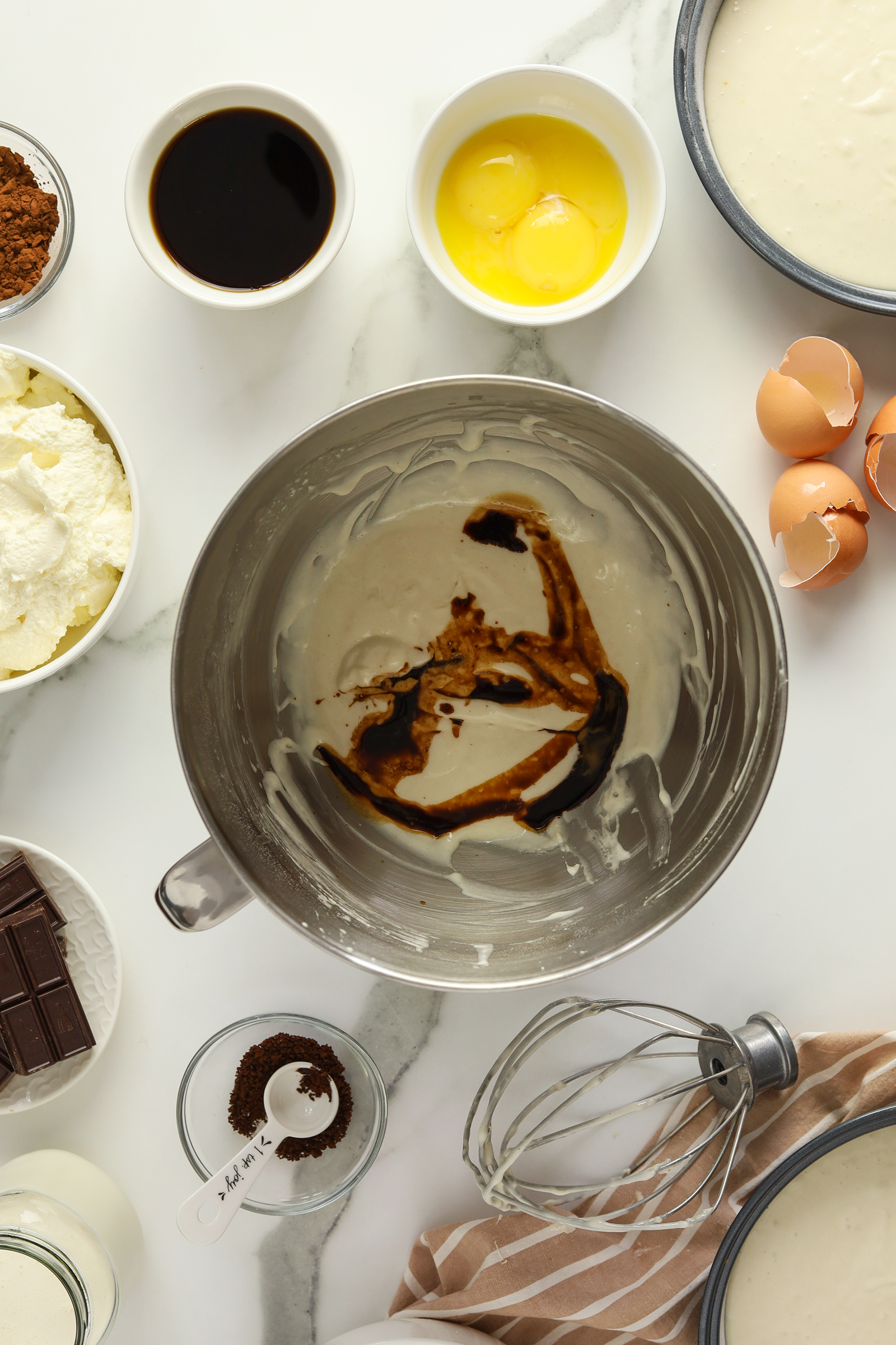 A mixing bowl with cake batter and vanilla extract sits on a white surface surrounded by eggs, ricotta cheese, cocoa, chocolate, coffee, and a whisk, ready for baking.