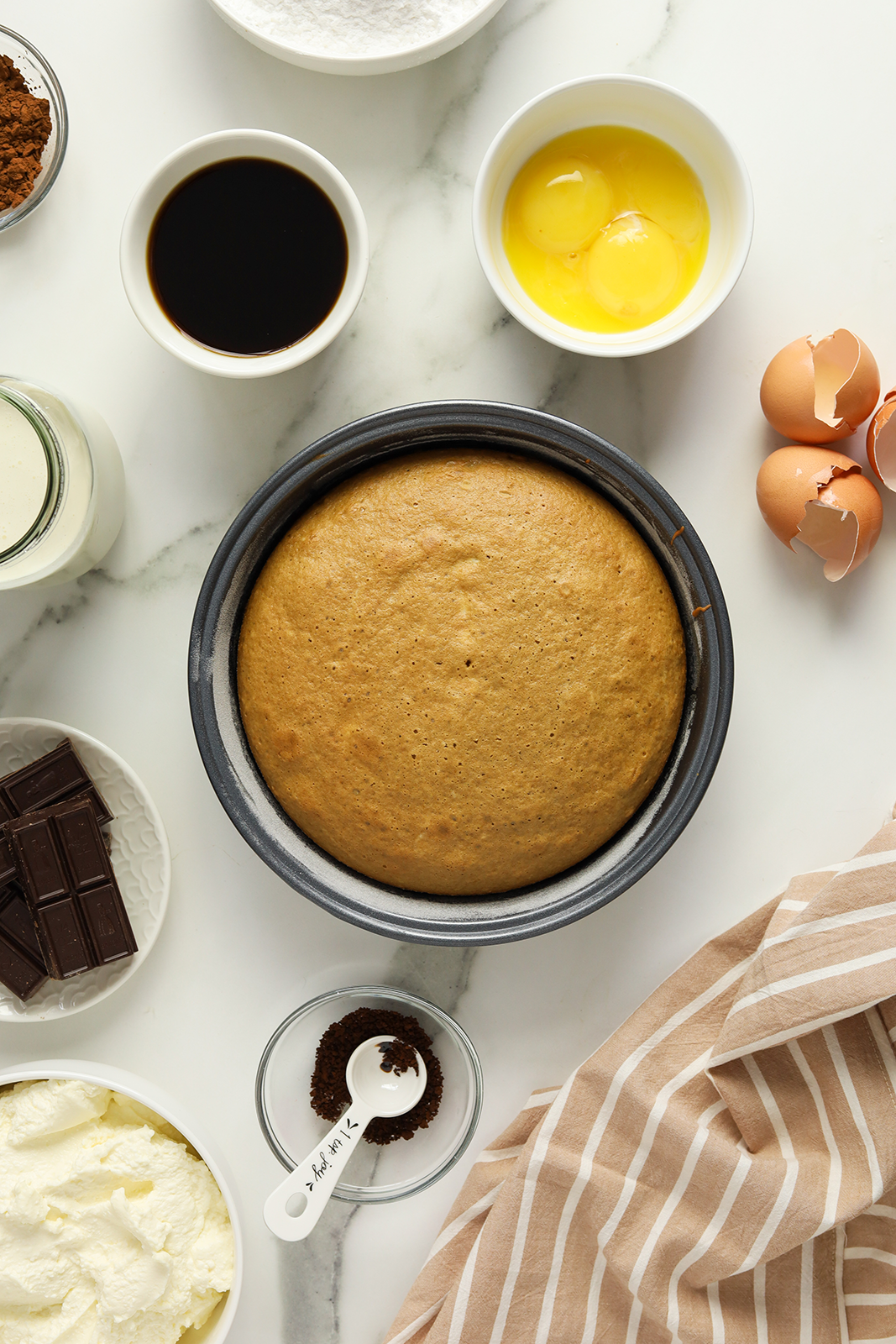 A round cake in a baking pan is surrounded by ingredients including eggs, chocolate, cream, cocoa, coffee, milk, and measuring spoons on a marble countertop with a striped cloth.