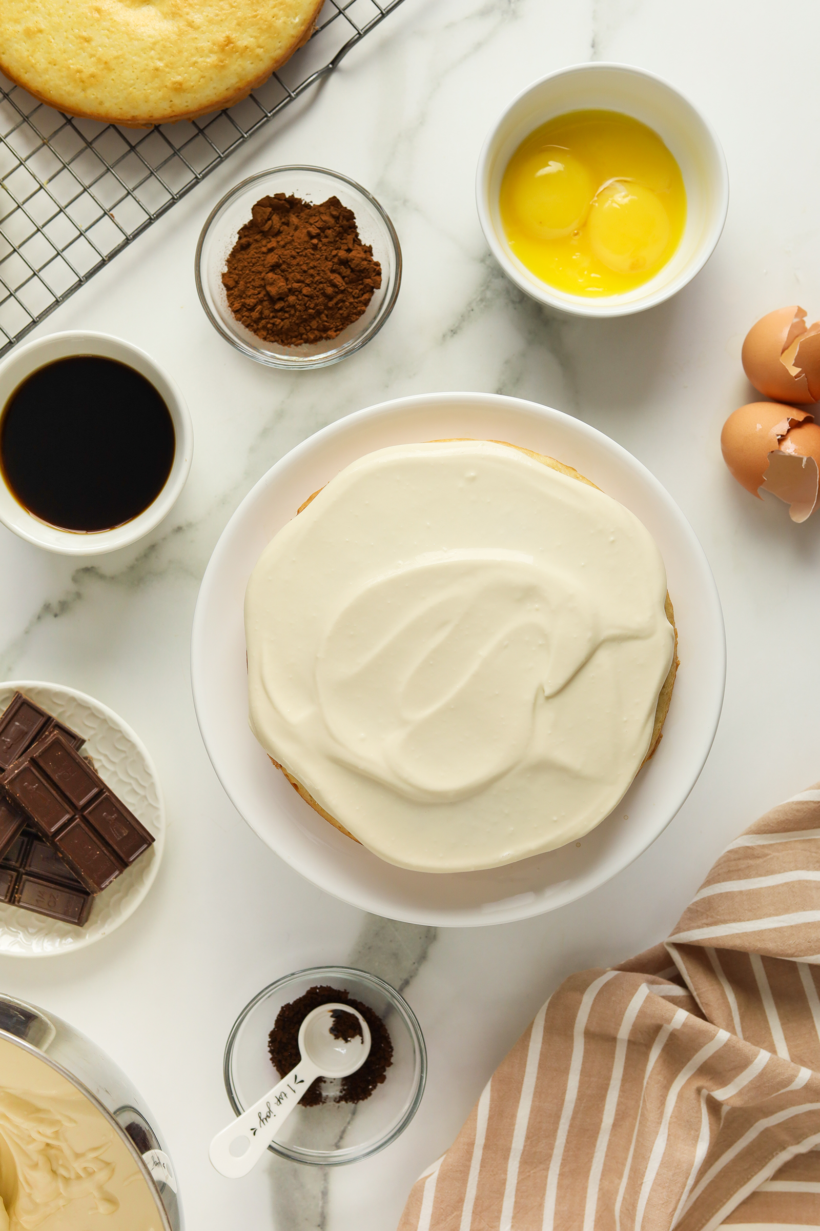 A cake with white frosting sits on a plate surrounded by baking ingredients: eggs, cocoa powder, chocolate, vanilla extract, and measuring spoons on a marble countertop with a striped cloth and a cooling rack.