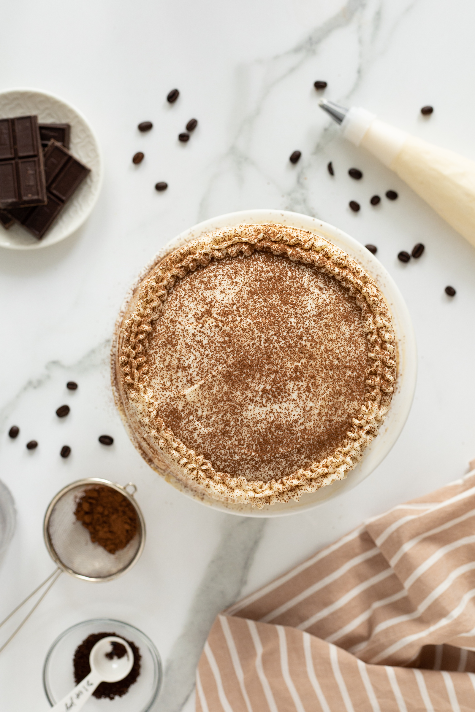 A top view of a tiramisu cake dusted with cocoa powder, decorated with piped cream along the edge. Surrounding it are coffee beans, chocolate pieces, a piping bag, instant coffee, and a striped cloth on a marble surface.