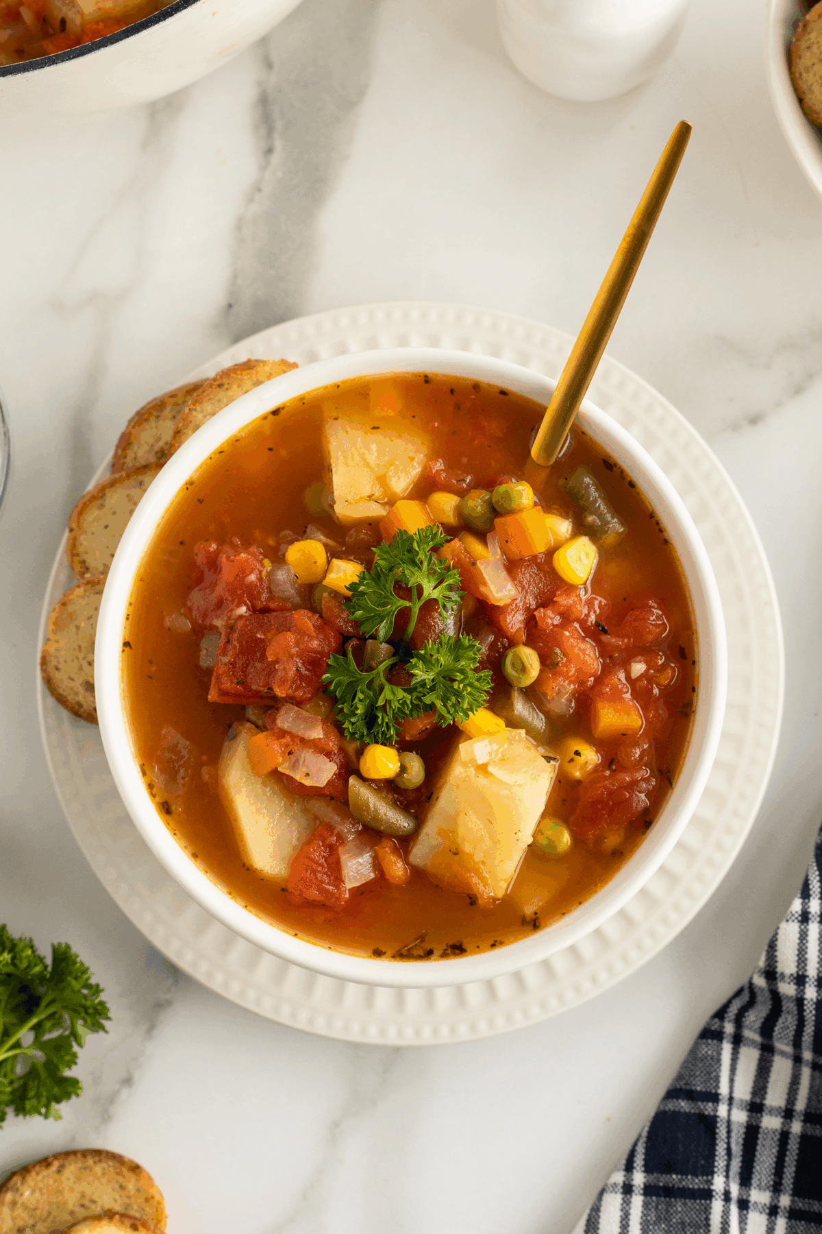 A bowl of vegetable soup with potatoes, tomatoes, corn, peas, and green beans, garnished with parsley, sits on a white plate with a spoon and sliced bread nearby.