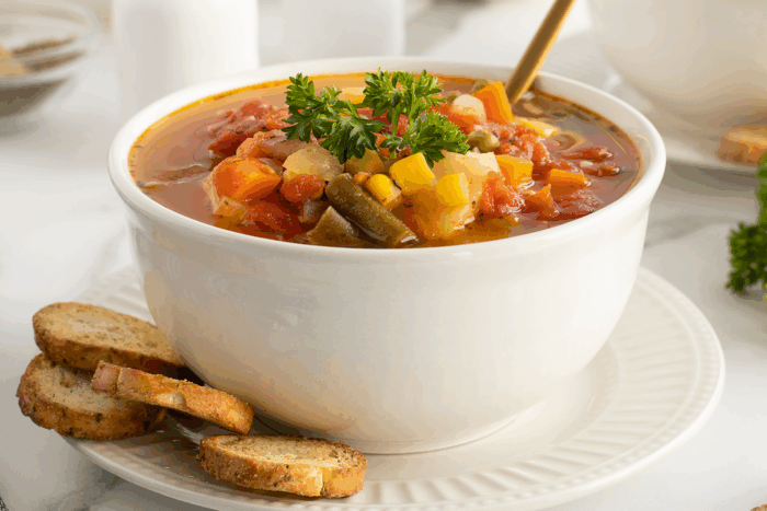 A white bowl filled with vegetable soup, topped with parsley, sits on a white plate with three slices of toasted bread on the side. A spoon is placed inside the bowl.