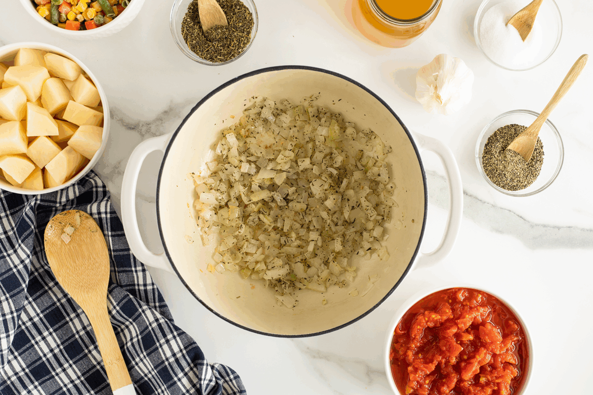 A white pot with sautéed onions and herbs sits on a marble counter, surrounded by bowls of chopped potatoes, canned tomatoes, mixed vegetables, spices, garlic, a wooden spoon, and a blue plaid towel.