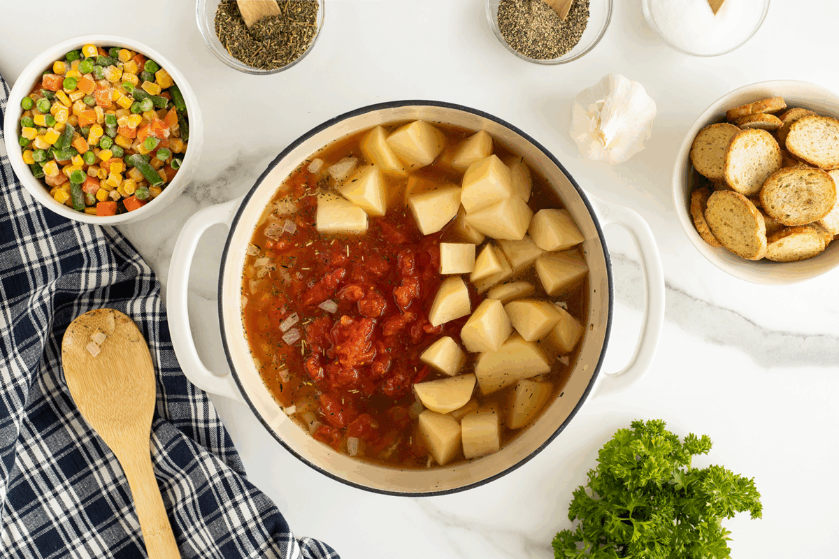 A pot of soup with diced potatoes and tomatoes sits on a counter, surrounded by a wooden spoon, a plaid towel, sliced bread, mixed vegetables, herbs, garlic, and fresh parsley.