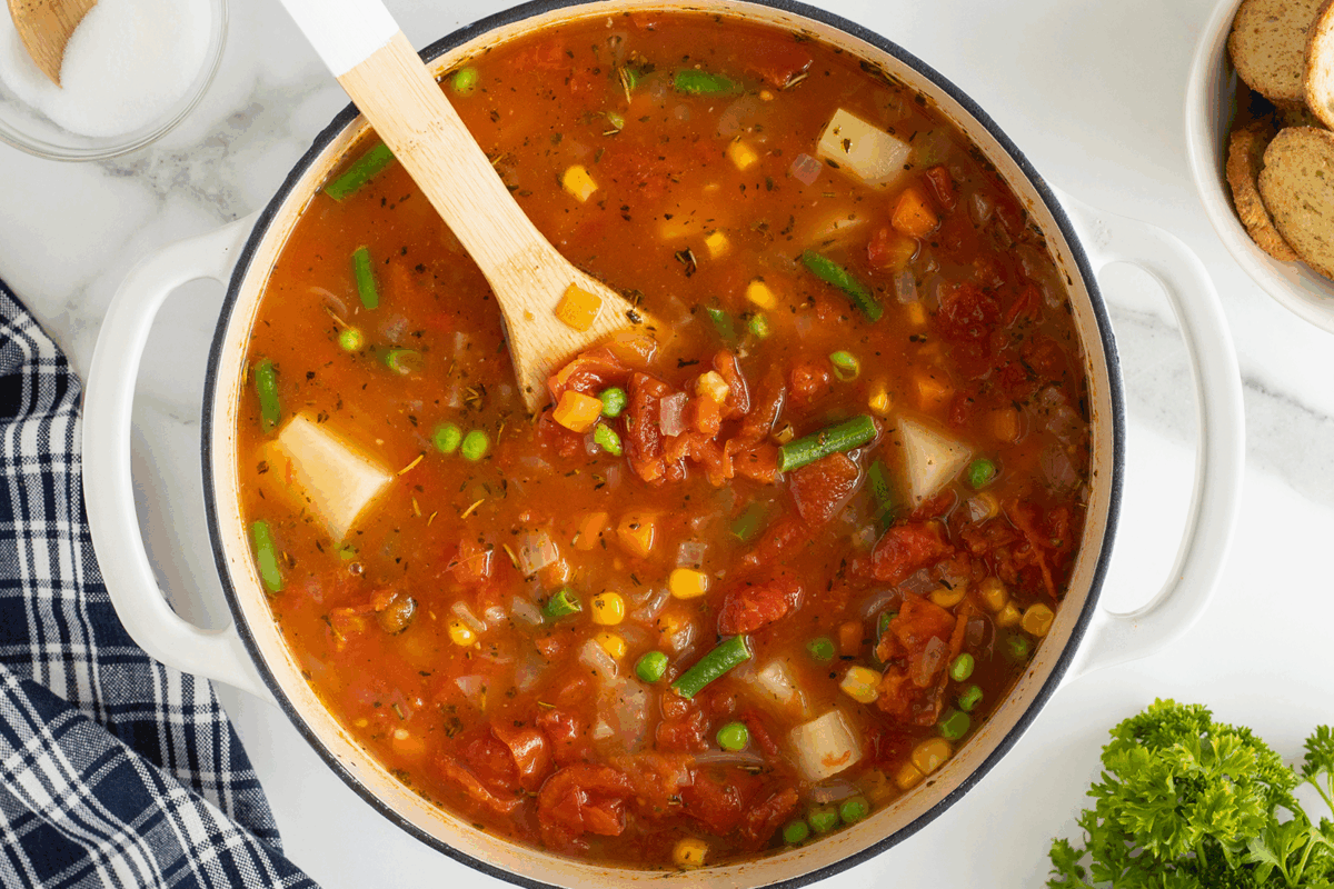 A pot of vegetable soup with tomatoes, potatoes, green beans, peas, and corn sits on a white surface. A wooden spoon rests in the pot. Nearby are a blue plaid napkin, fresh parsley, and a plate of bread slices.