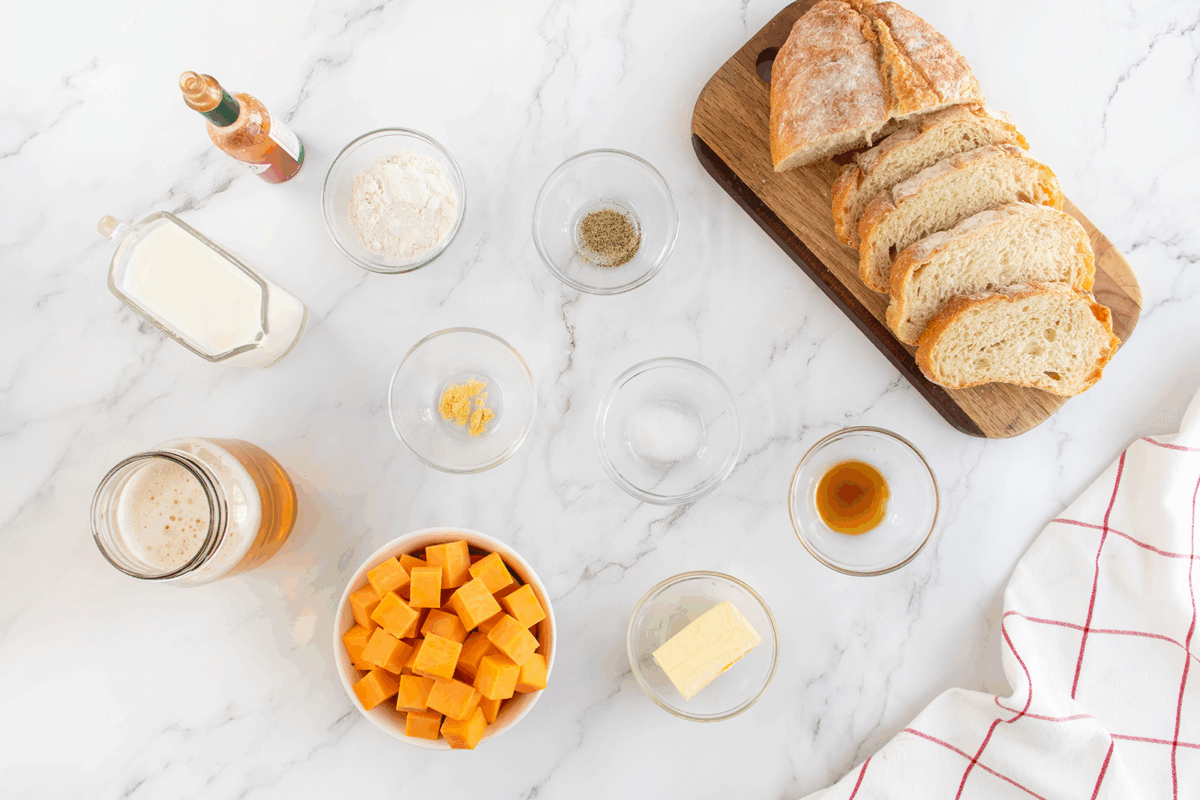 A marble countertop displays sliced bread on a wooden board, bowls of cubed cheese, butter, flour, seasonings, minced garlic, a glass of beer, milk, Worcestershire sauce, and a bottle of hot sauce.