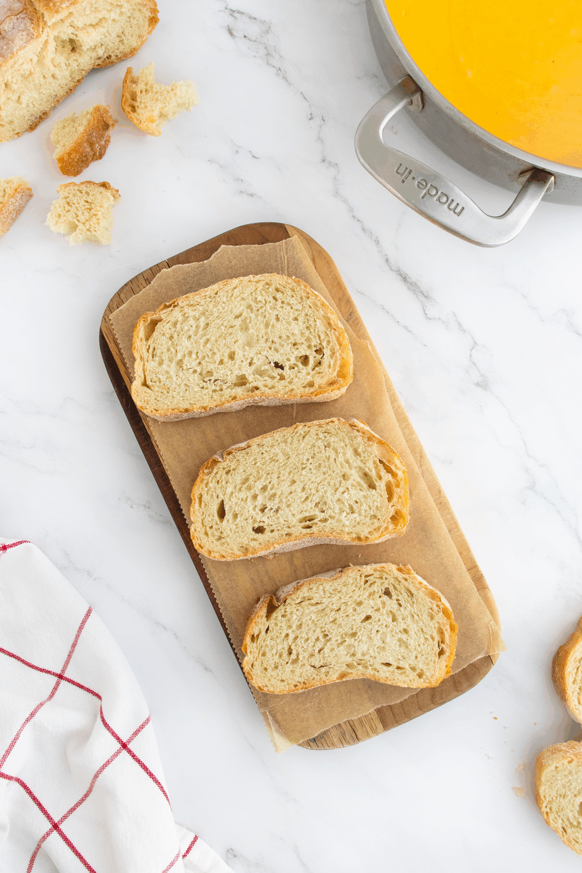 Three slices of rustic bread on a wooden board lined with parchment paper, surrounded by bread crumbs and a soup pot on a white marble surface. A red-and-white kitchen towel is partially visible in the corner.