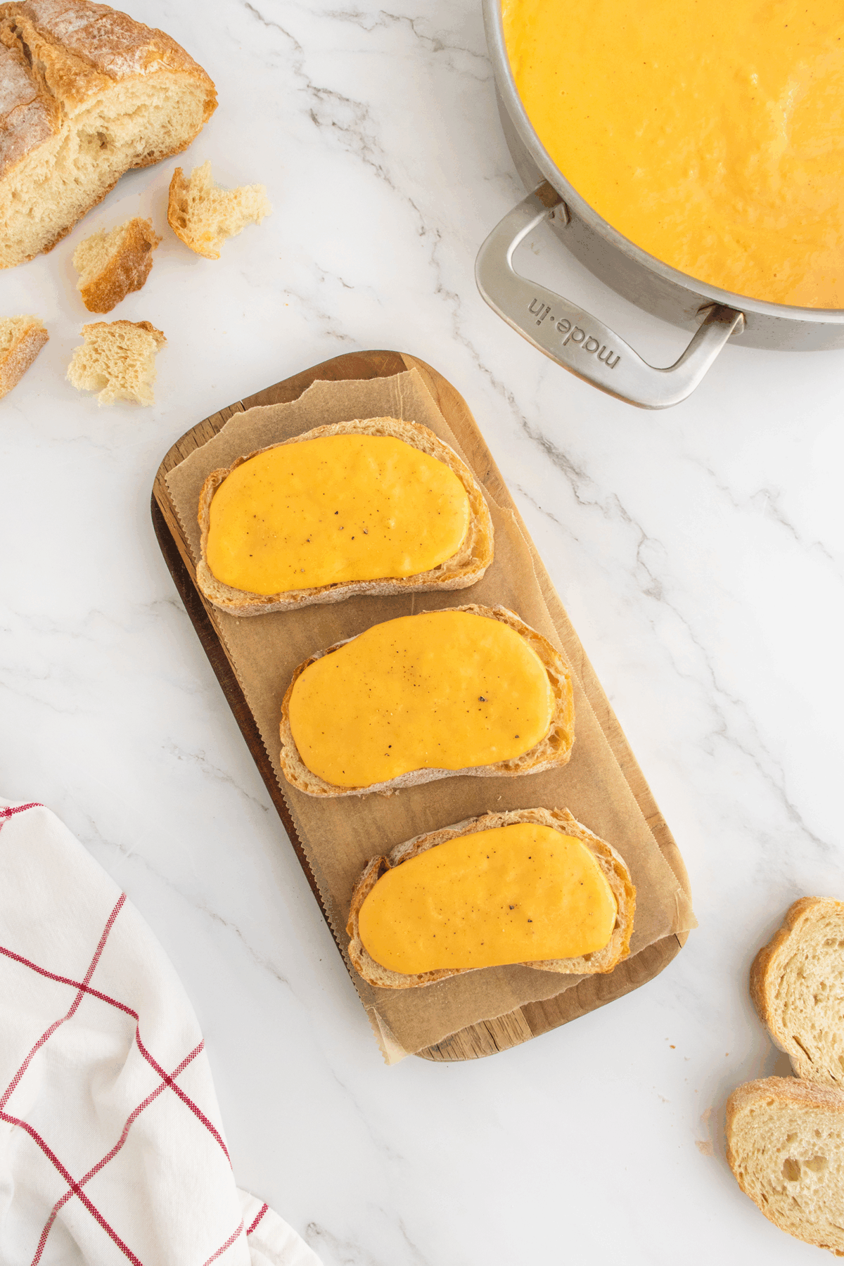 Three slices of bread are topped with melted cheese and placed on a parchment-lined wooden tray. A pot of cheese sauce, bread slices, and crumbs are visible on a white marble surface.