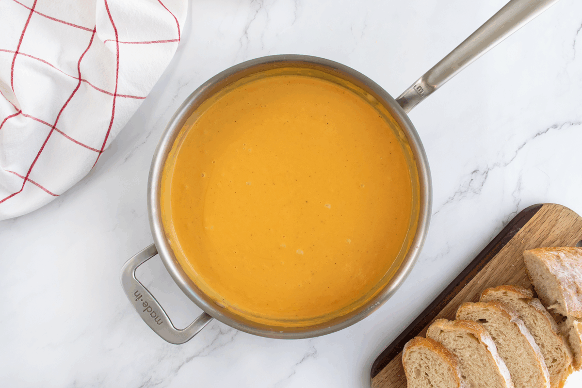 A saucepan filled with creamy orange soup sits on a marble surface next to a checkered kitchen towel and a wooden board with sliced bread.