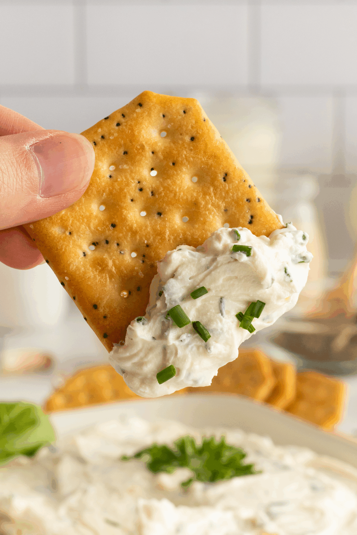 A hand holds a poppy seed cracker with a generous dollop of creamy, chive-topped dip above a bowl of dip, with more crackers blurred in the background.